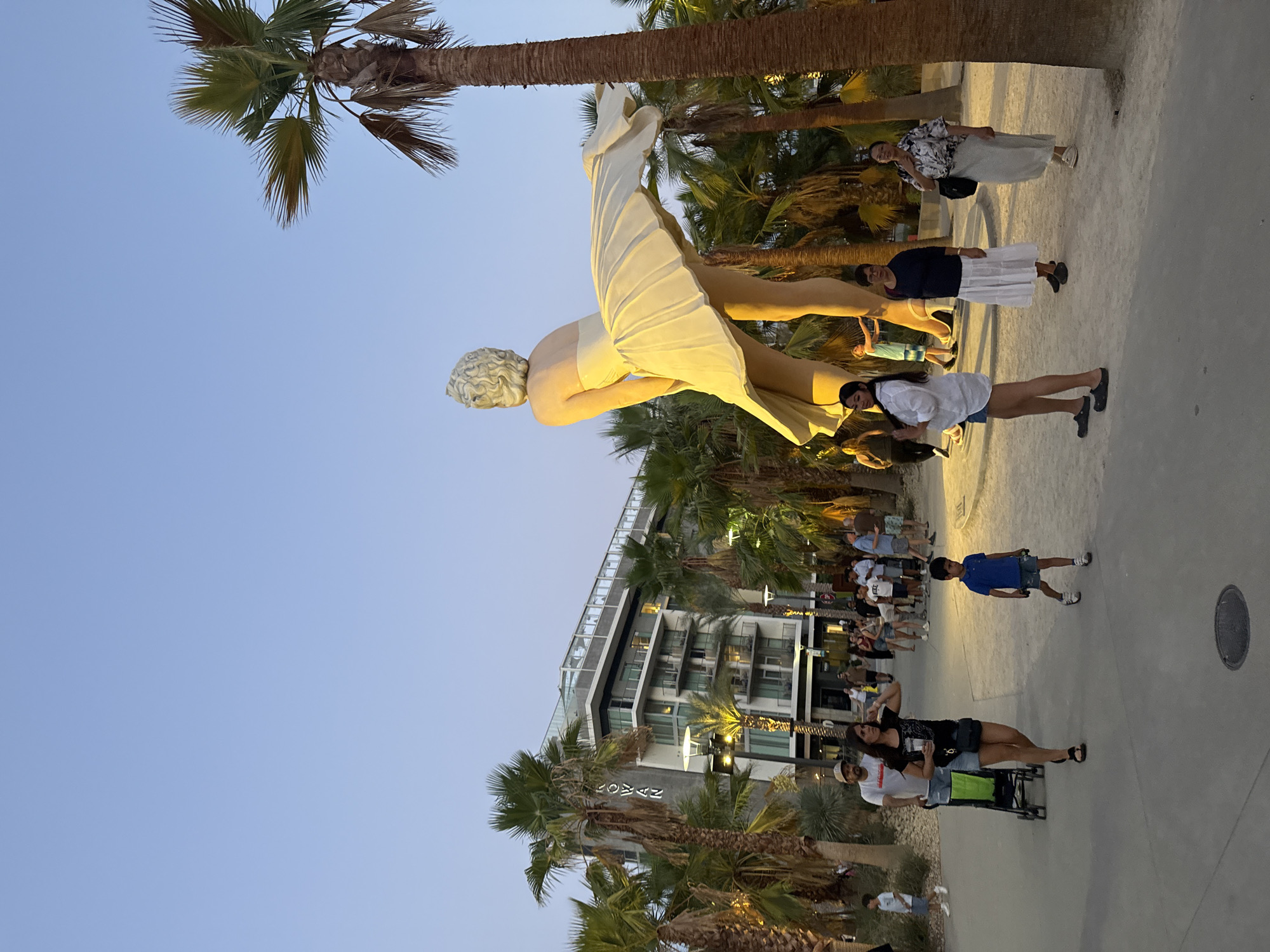 The image captures a public outdoor scene in Palm Springs, California, with a large statue of Marilyn Monroe from "The Seven Year Itch" as the central focus. The statue depicts Monroe in her iconic white dress, with the skirt billowing upwards. It is positioned amidst numerous palm trees and a modern building in the background, identified as the "ROWAN" hotel.
Several people are visible in the foreground and middle ground, engaged in various activities. A woman in a black top and denim shorts walks with a stroller, accompanied by a man wearing a white t-shirt and a cap. To the right, a young boy in a blue polo shirt and shorts stands looking forward. Further to the right, a woman in a white shirt and denim shorts poses, and two women in long skirts and tops are also present. Other individuals are seen in the background, some walking, others standing and observing the statue.
The environment is characterized by sandy ground, paved walkways, and lush palm trees, suggesting a resort or entertainment district. The lighting indicates it is late afternoon or early evening, with a clear sky and warm illumination. The building in the background is a multi-story modern structure with balconies and visible windows, suggesting it is a hotel or residential complex. The overall atmosphere appears relaxed and leisurely.