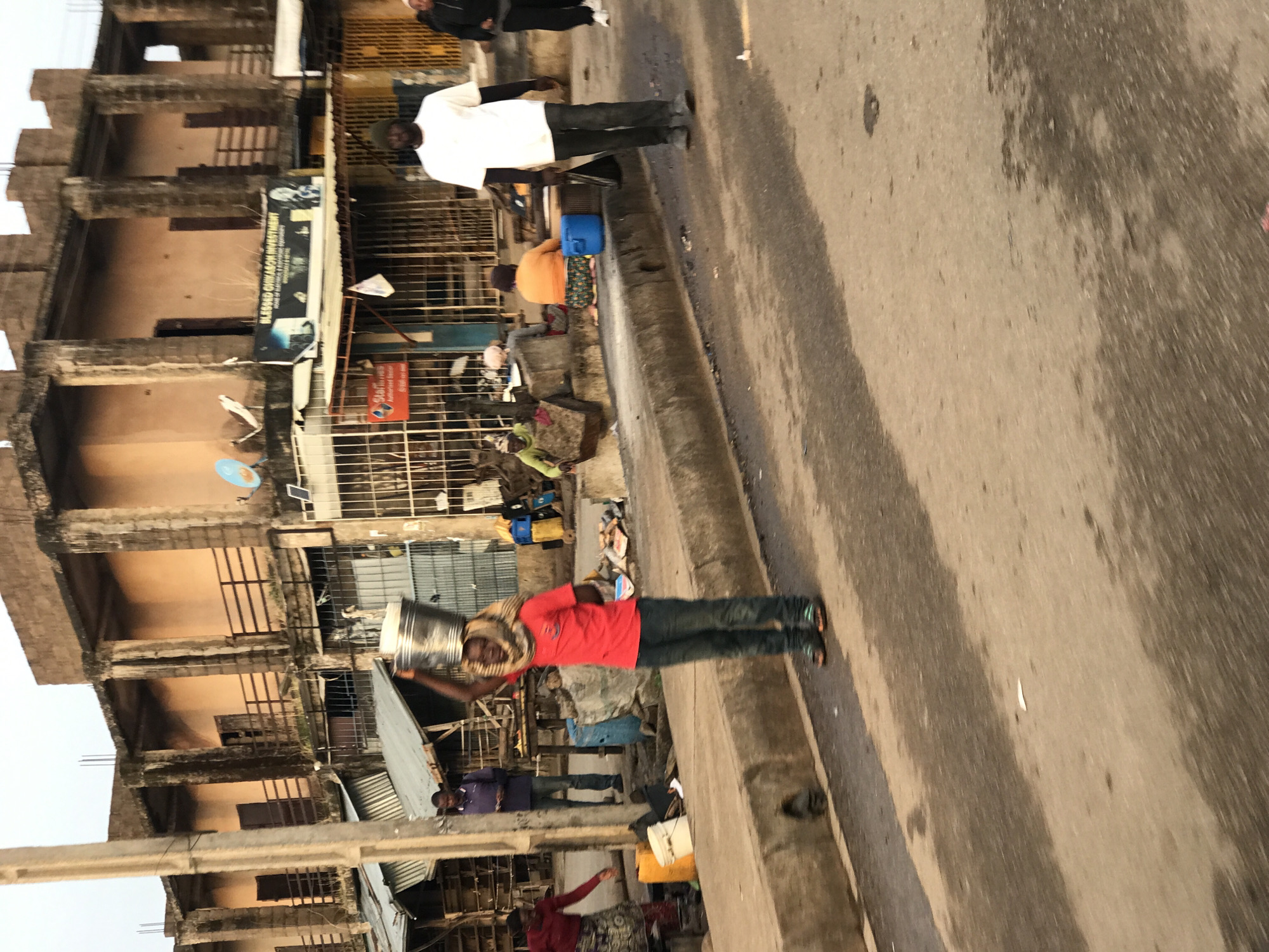 The image depicts a street scene in Kabong, Nigeria. In the foreground, a person in a red shirt and blue jeans is walking with a metal bucket balanced on their head, wrapped in a cloth scarf. To the right, a man in a white t-shirt and dark pants is walking away from the camera, and another person in dark clothing is walking towards the right. In the background, there is an old, unfinished building with visible concrete columns and balconies. Below the building, market stalls with corrugated metal roofs and metal grates are visible. Several people are seen sitting or standing near the stalls, some engaged in what appears to be market activities. The street is paved with asphalt and has a concrete curb. Various objects, including yellow and blue plastic containers, are scattered along the roadside and in front of the stalls. The overall environment appears to be an outdoor market area within a built-up urban setting.