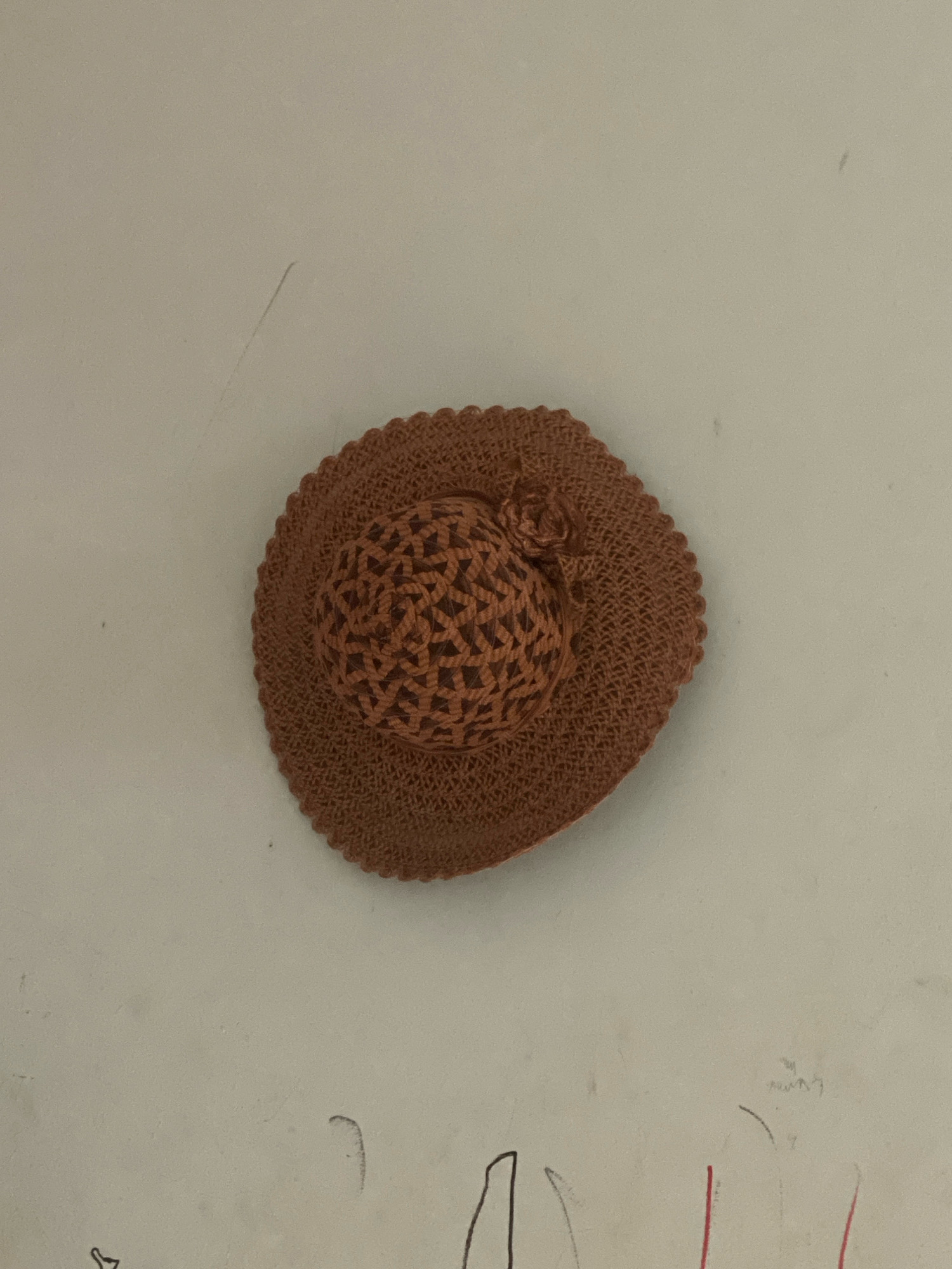 A brown, crocheted straw hat with a woven dome and a decorative rosette is positioned centrally against a light-colored, textured background. The background appears to be a wall with faint scuff marks and some rudimentary crayon drawings at the bottom, suggesting a domestic or possibly child-friendly environment. The lighting is neutral, casting no significant shadows. The specific location in Batam, Indonesia, cannot be determined from the image alone.