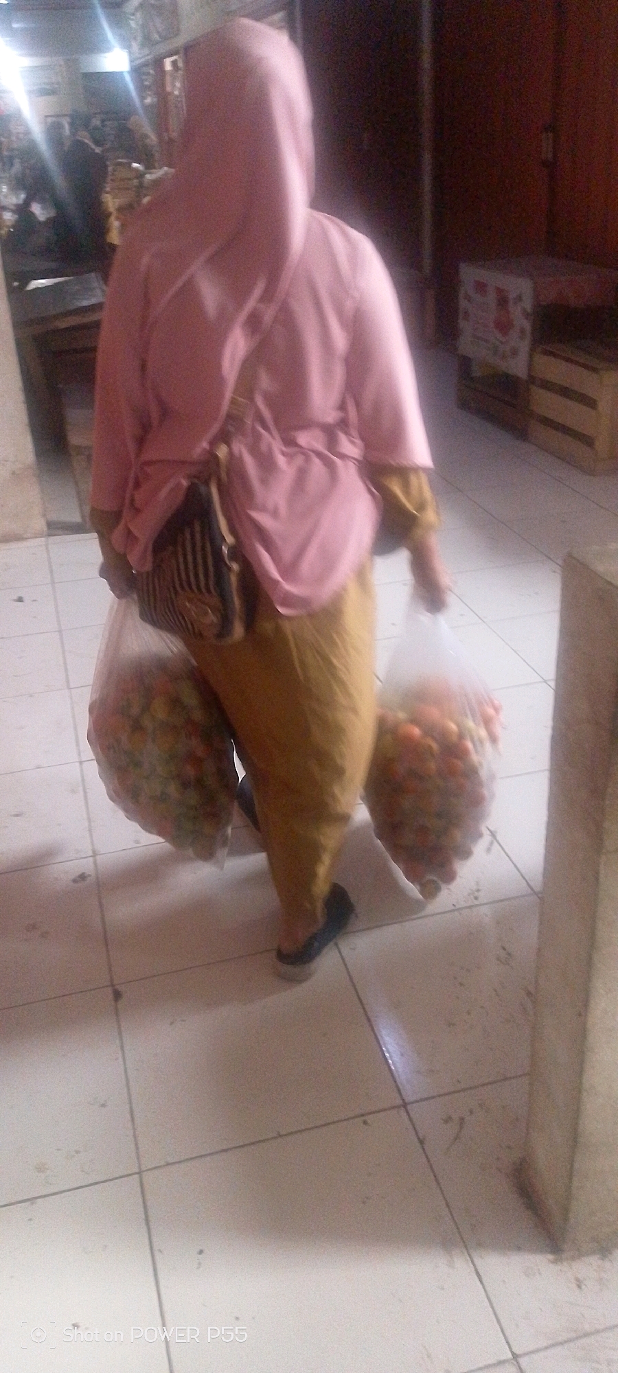 A person wearing a pink hijab and a yellow skirt walks while carrying two plastic bags filled with fruits. The person also has a shoulder bag. The environment appears to be an indoor market with tiled floors and some wooden crates in the background.