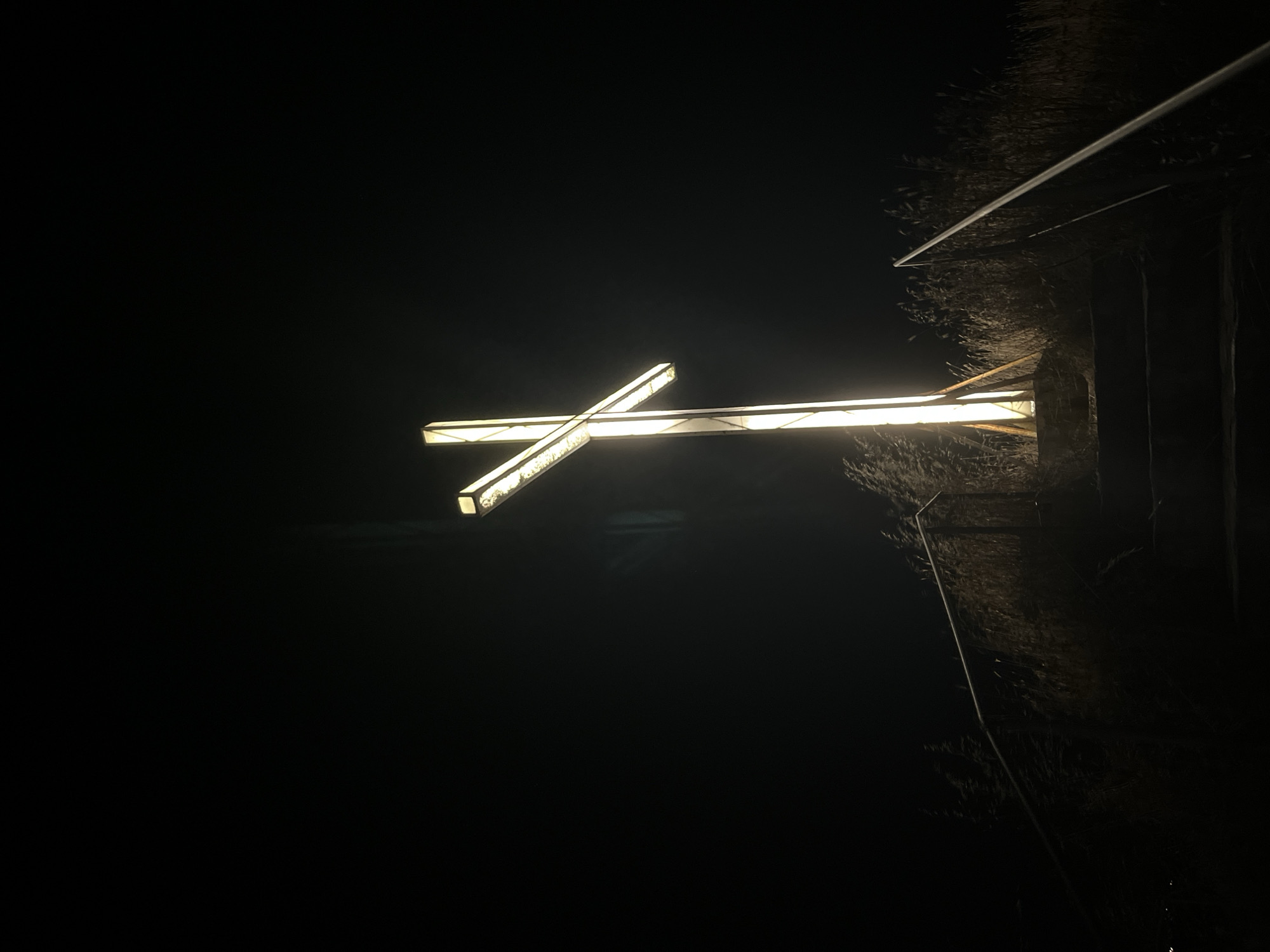 The image depicts a lit cross standing tall against a dark, night sky. The cross is illuminated with a bright, warm light, casting a glow around its structure. The bottom of the cross appears to be anchored on a raised platform, with a set of stairs leading up to it. These stairs are bordered by metal railings on both sides. Tall, dry grass or reeds surround the base of the cross and the stairs, suggesting a natural or somewhat untamed environment. The overall scene is one of solemnity and prominence, with the glowing cross being the central focal point against the deep darkness of the night. There are no people or animals visible in the frame. The location is identified as being in Lozenets, Bulgaria.