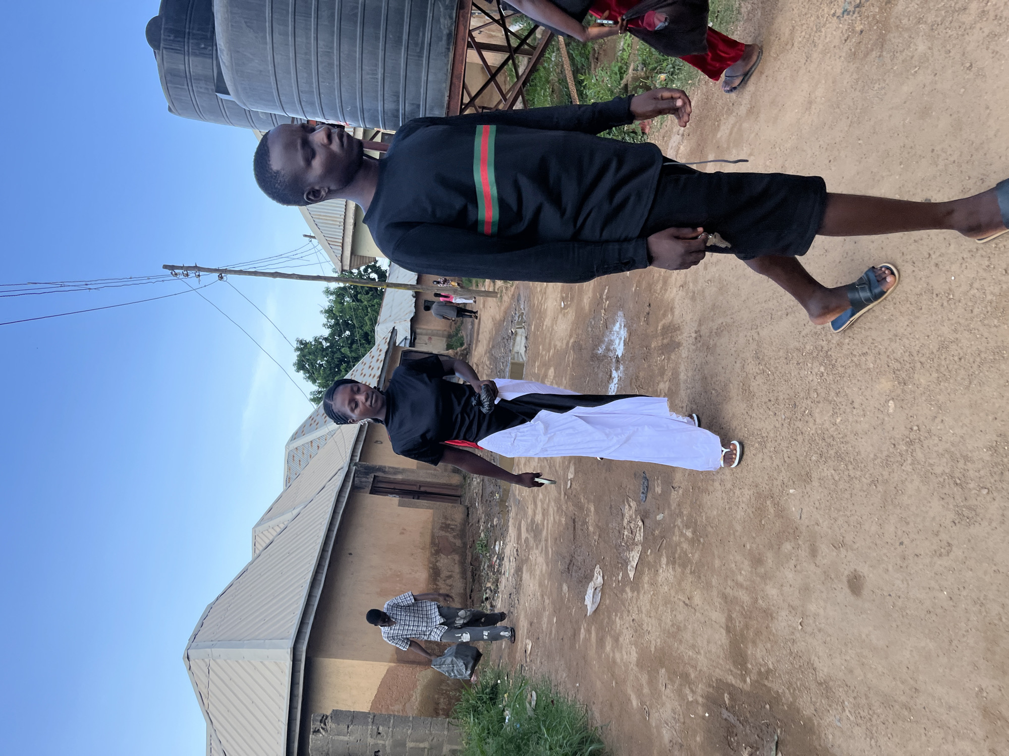 In the city of Rafin Par, Nigeria, a young man, predominantly in the foreground and right of the frame, walks towards the viewer, his gaze directed slightly off-camera to the right. He wears a black long-sleeved shirt with red and green horizontal stripes across the chest and black shorts. His left hand is raised, holding a set of keys, and he wears blue and black sandals. To the left of the frame, a young woman walks away from the viewer, her back turned. She is dressed in a black top and a long white skirt, with a black sash. She holds a black object in her right hand and wears white sandals. Behind her, another individual in a plaid shirt walks away from the viewer, carrying a large black plastic bag in his right hand. In the background, several single-story, light brown buildings with corrugated metal roofs line a dirt road. Power lines stretch across the sky. To the right of the image, large black water tanks stand on a metal structure. The ground is a dusty, reddish-brown dirt path, with scattered debris and small puddles of water. The sky is a clear blue with a few faint clouds.