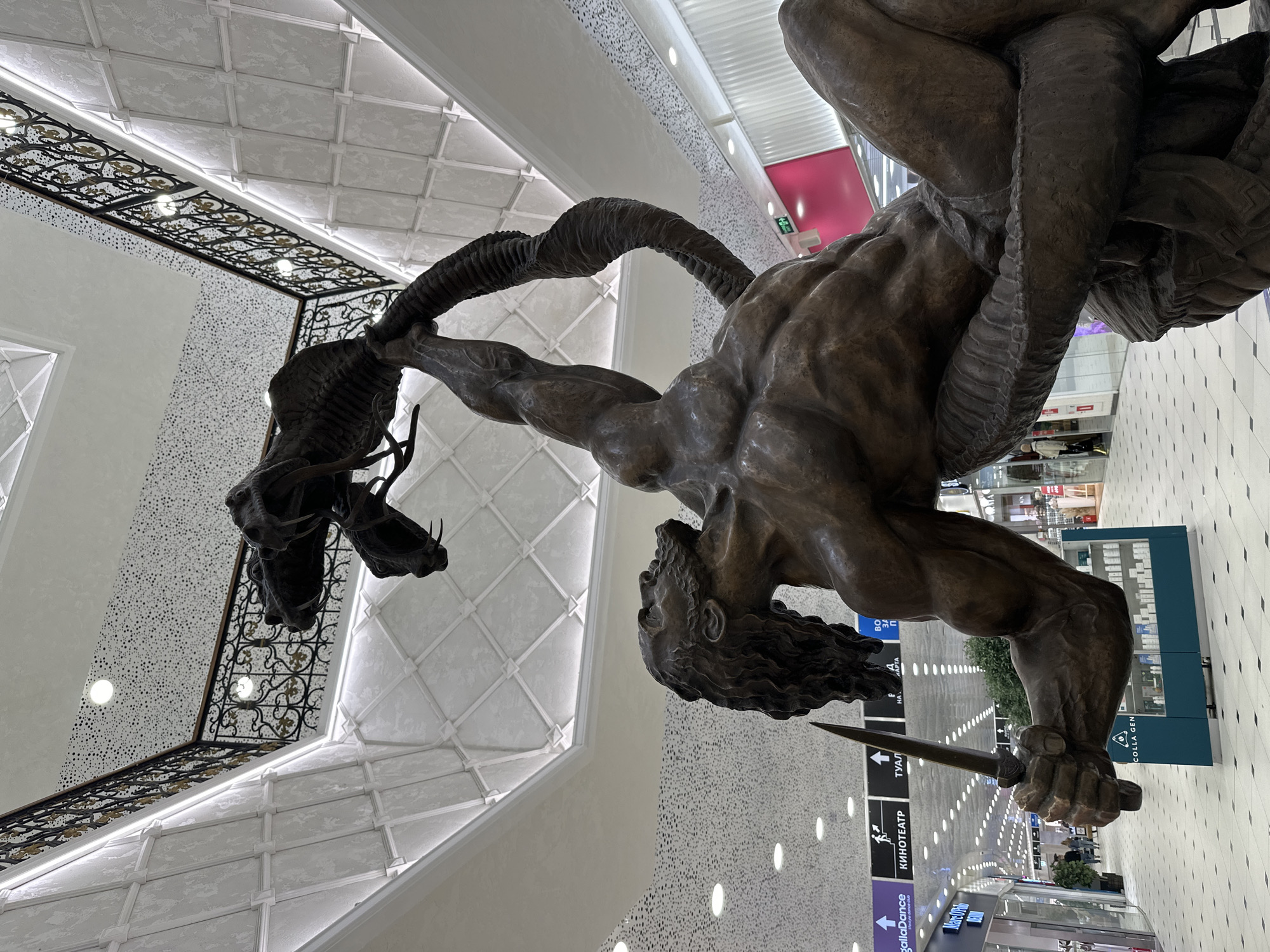 A large, detailed bronze statue depicting a muscular man in a dynamic struggle with a serpentine or dragon-like creature is the central subject of the image. The man, with flowing, curly hair, is shown actively restraining the creature with one arm around its neck or horns, while his other hand grips a sword, poised for action. The creature is characterized by prominent horns or antlers and a textured, scaled body, conveying a sense of ferocity.
The statue is situated indoors, within a spacious and brightly lit modern architectural setting, strongly resembling a multi-story shopping mall or a large public entertainment complex. The high ceiling features decorative coffered panels with integrated linear lighting. Ornate black metal railings with gold accents are visible on an upper level, suggesting balconies or additional floors. The walls display varied finishes, including a patterned surface with numerous small white dots and a striking bright pink section in the background. The flooring is composed of light-colored tiles arranged in a grid pattern. Rows of recessed spotlights line the ceiling of a long corridor stretching behind the statue, indicating a busy thoroughfare.
The scene captures a dramatic moment of mythological or heroic combat, set within a sleek, contemporary public space. The artificial lighting ensures a bright and inviting atmosphere, typical of commercial venues.
Visible text on various signs in the background includes:
* "КИНОТЕАТР" (Kinoteatr), which translates to "Cinema."
* "ТУАЛЕТ" (Tualet), meaning "Toilet."
* "COLLAGEN," likely a brand or product name.
Additional partial or blurry Russian text, such as "ВО ЗДАРОВЬЯ" and "АРТА," can also be seen on other signs.