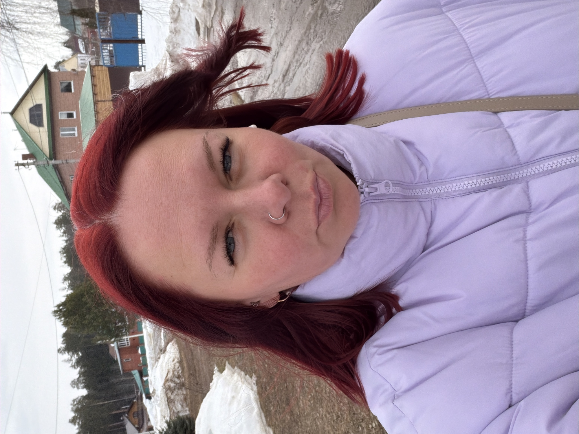 This is an outdoor portrait (selfie) of an adult female in Chaikovsky, Russia, taken during what appears to be late winter or early spring conditions. The female, positioned centrally and occupying the majority of the frame, has long, dyed red hair, blue eyes, and light skin. Her hair is slightly wind-blown on her right side (viewer's right). She wears a silver hoop septum ring, a small gold hoop earring with a star charm in her left ear, and a white stud earring in her right ear. Her expression is direct towards the camera with a slight pout. She is dressed in a light lavender/lilac colored puffy winter jacket, zipped up to her neck. A light beige or tan strap from a bag is visible over her right shoulder.
The background depicts a residential or semi-rural environment. Patches of melting snow are visible on uneven, brownish ground, suggesting a dirt path or unpaved area. In the middle distance, several structures are present: a two-story brick house with a prominent green gabled roof and a light-colored dormer, and another brick house with a dark green roof and a bay window or balcony. Further left, partially obscured by trees, is a brown wooden house with a light roof. A blue metal gate and fence are visible on the right side of the background. Various trees, including coniferous evergreens and bare deciduous trees, populate the landscape. Overhead, power lines traverse the upper portion of the frame against an overcast, light gray sky.