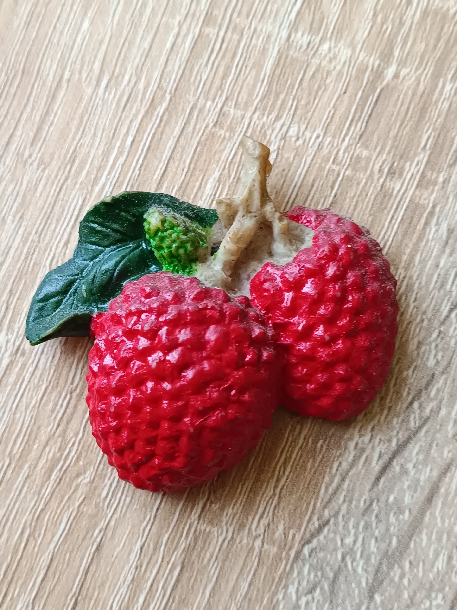 A decorative figurine of two red lychees with green leaves and a stem rests on a light brown wooden surface. The lychees have a textured, bumpy surface and are split open, revealing a white interior. The leaves are dark green and detailed, and the stem is light brown and twig-like. The background surface appears to be a laminate or wood with visible grain lines running diagonally. The scene is lit from above and to the right, casting subtle shadows. There are no people or any other objects present in the image. The image is a close-up shot, focusing entirely on the lychee figurine and the surface it rests on. This scene does not have any relation to Tebras, Latvia.