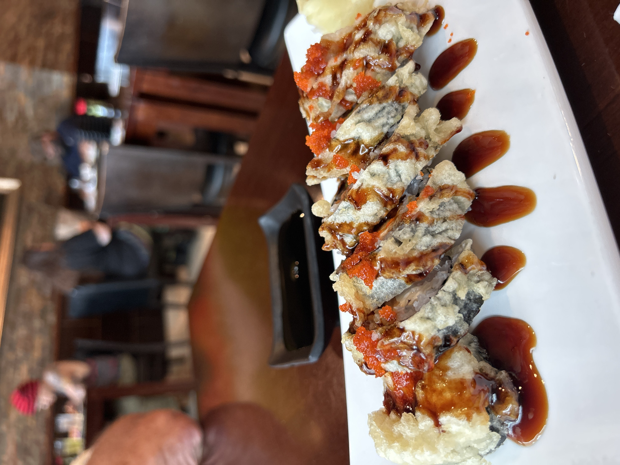 A close-up, eye-level shot captures a plate of eight pieces of fried sushi rolls artfully arranged on a white rectangular plate. The rolls are generously drizzled with a dark, glossy sauce, and a scattering of vibrant orange roe adorns the tops of several pieces. A small black dish of soy sauce sits behind the sushi, and a sliver of pickled ginger is visible on the right side of the plate. The background is blurred, showing the interior of a restaurant with wooden tables and chairs, and indistinct figures of patrons seated and dining. The lighting appears to be ambient restaurant lighting.