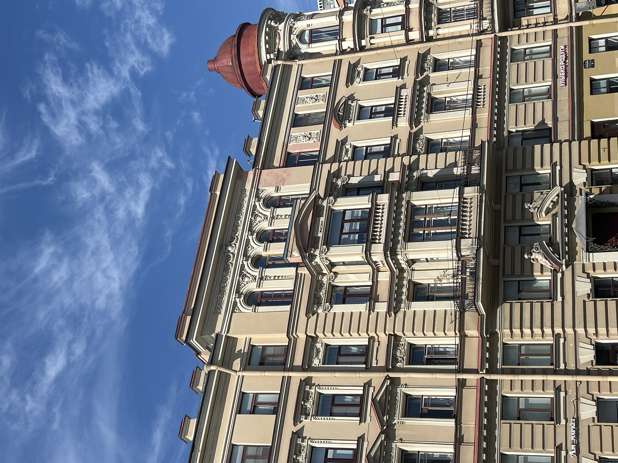 This is a low-angle, close-up shot of a building's facade against a bright blue sky with wispy white clouds. The building appears to be made of light-colored stone with intricate architectural details. Several stories of windows, some with dark frames and others with lighter frames, are visible. The building exhibits ornate carvings and decorative elements, including balconies and what appears to be a red dome or cupola at the top left. The sunlight suggests it is daytime, and the clear sky indicates fair weather. A vertical sign with Cyrillic text, reading "УЛЫБКА РАДУГИ," is visible on the facade of the building. The scene is an urban setting, focusing on the architectural style of the building. There are no people or clear indications of a specific event taking place.