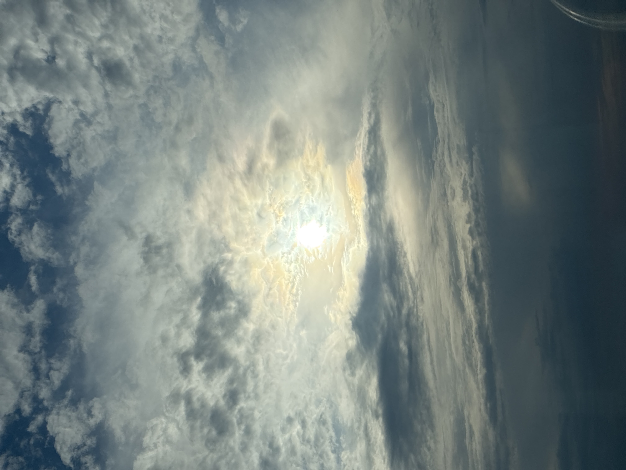 The image depicts a cloudy sky with the sun peeking through. The sun is centrally located, casting a bright glow through a break in the clouds. The clouds are a mix of white and grey, with some appearing darker and denser. The overall impression is of a dynamic sky, possibly taken from an airplane due to the slight curvature at the bottom right, suggesting a window. No specific city or location is discernible.
