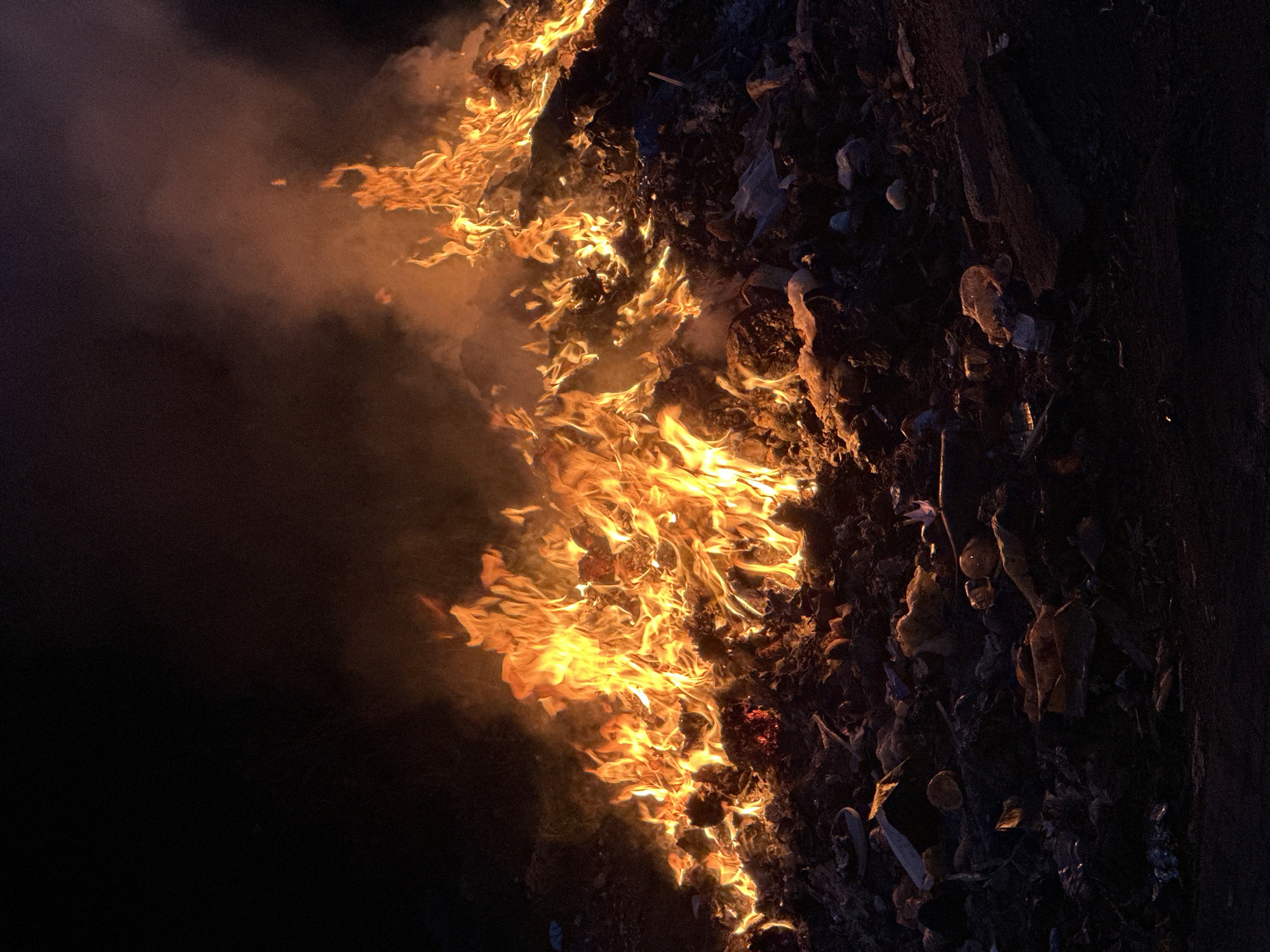 The image depicts a fire engulfing debris or trash in an outdoor setting. The flames are prominent, displaying a range of yellow and orange hues, and are concentrated along a vertical line on the left side of the frame. Dark, unidentifiable objects are visible to the right of the flames. Smoke is present, emanating from the fire. The background is dark, with very little visible detail. There is no visible evidence indicating the city of Bac Nha Trang Ward, Vietnam. There are no people present in the image.