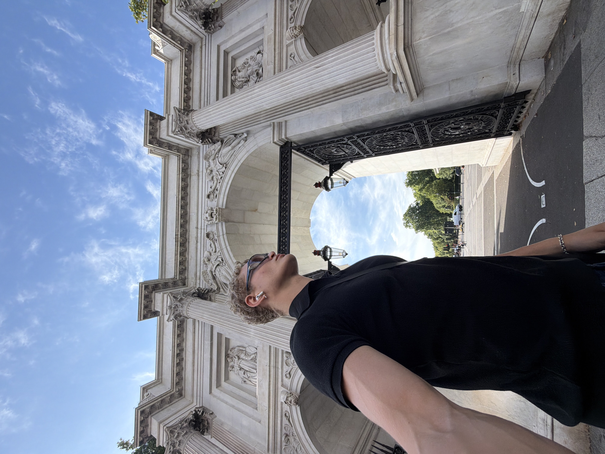 A full-body, slightly tilted portrait shows a young, blonde, white male in his early twenties standing in front of the Marble Arch in London, United Kingdom. He is wearing a black polo shirt, dark sunglasses, and white AirPods. The Marble Arch, a triumphal arch made of white marble, is visible behind him. The arch features intricate carvings and columns. The sky is blue with scattered white clouds. In the background, trees and a street with traffic can be seen. The man is looking to his right, with his left arm extended towards the camera.