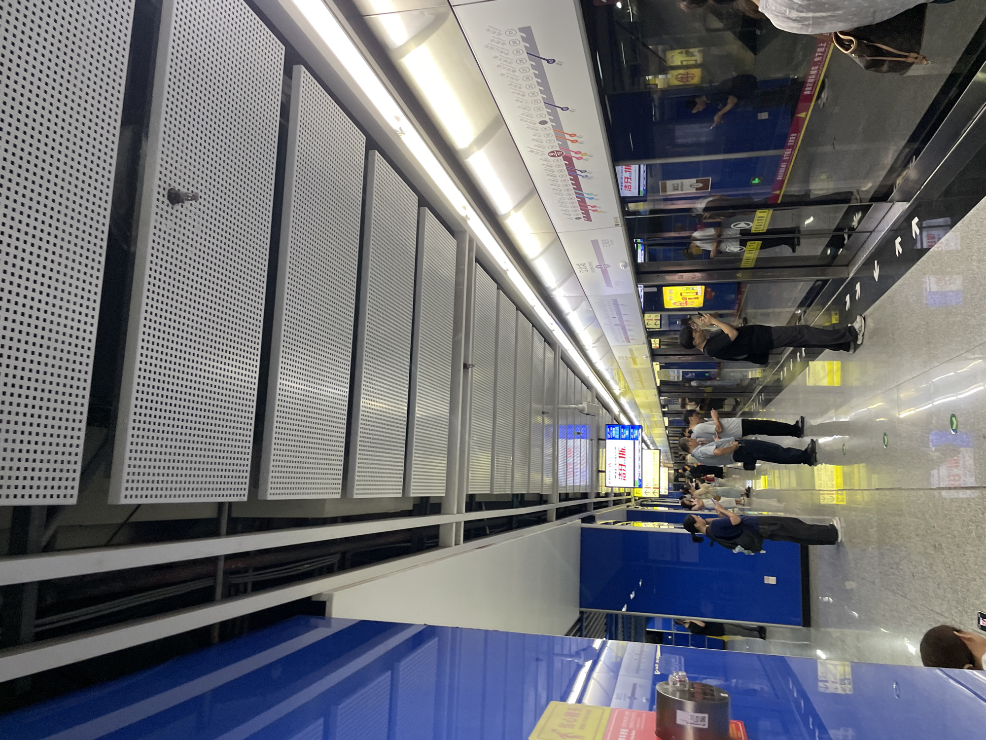 An indoor, eye-level, wide shot shows a modern, well-lit subway station platform. The platform is made of speckled grey tiles with white arrows indicating walking direction. On the left, a tall blue wall features a reflective surface. Above, a white, perforated ceiling is divided into rectangular panels, with exposed conduits and lighting fixtures.
To the right, a train is stopped at the platform. The train doors are glass, reflecting the station and passengers. Above the doors, a long, illuminated display shows a subway map with station names and line colors. Passengers are standing on the platform, many looking at their phones. Several individuals are visible, including a woman in black with a backpack, a man in a grey shirt and black pants, and another man in a black t-shirt and grey pants. The overall atmosphere is orderly and typical of a public transportation hub. The city is Unknown, Unknown.