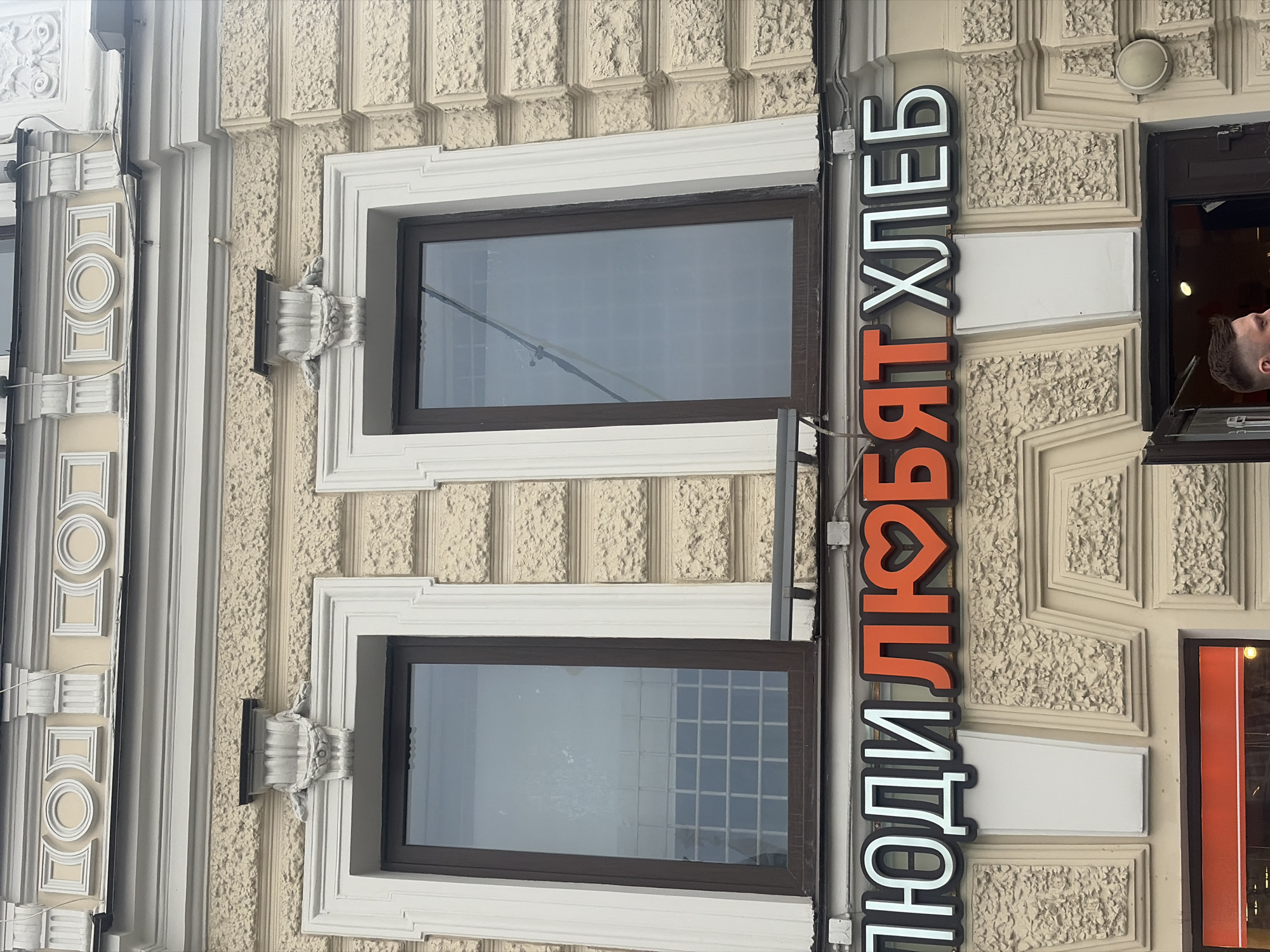 The image depicts a section of a light-colored building facade in Saint Petersburg, Russia, featuring architectural ornamentation and commercial signage. The building's exterior is predominantly beige with a textured, rusticated finish on the lower visible sections and smoother, white classical moldings above. Two tall, rectangular windows with dark brown frames are centrally positioned, reflecting an overcast sky and internal grid patterns. Above these windows, the facade includes decorative white elements such as cornices, pilasters, a frieze with repeating circular and square motifs, and stylized garlands positioned directly above each window.
Below the windows, a prominent, illuminated channel letter sign displays the Russian text "ЛЮДИ ЛЮБЯТ ХЛЕБ" (People Love Bread). The words "ЛЮДИ" and "ХЛЕБ" are white with black outlines, while "ЛЮБЯТ" is orange with a black outline, and the letter 'Б' is stylized as a red heart. Electrical conduits and junction boxes are visible on the facade near the sign and above the windows.
In the bottom right corner, a dark-colored door is open, revealing a glimpse of a lit interior. A male individual, visible from the head to the upper shoulder, with short dark hair and a light-colored shirt, is positioned near the doorway, appearing to enter or exit. He is looking slightly to his left. A round, white exterior light fixture is mounted on the wall to the right of the entrance. The overall scene is an urban outdoor setting.