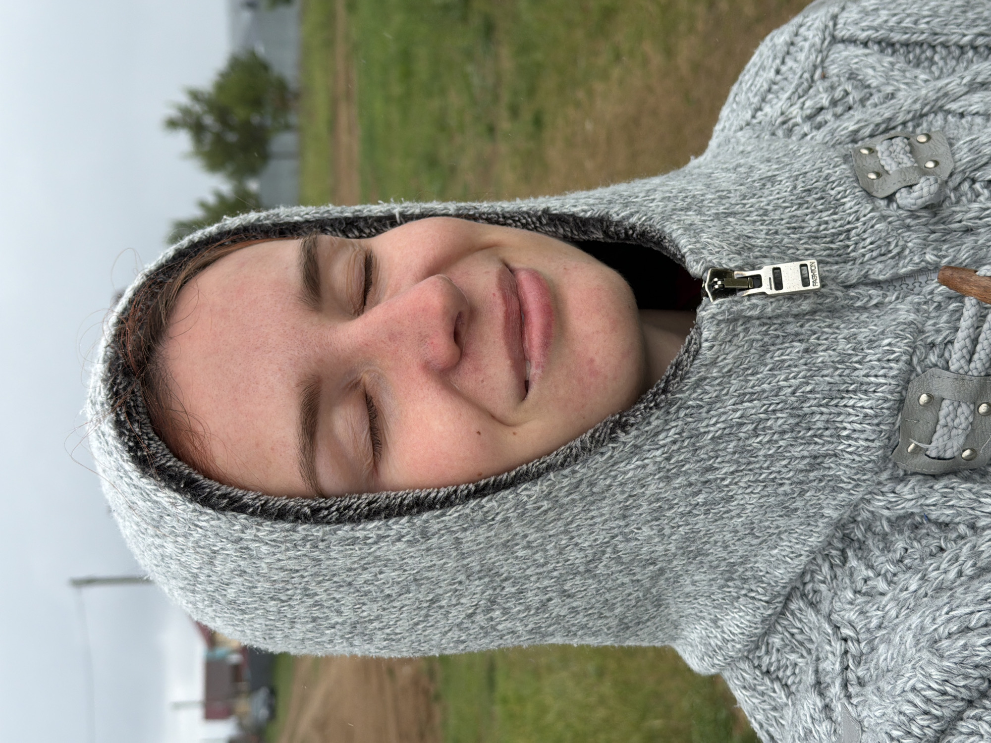 A young woman is wearing a gray knit hooded sweater zipped up to her neck outdoors. Her eyes are closed and she is smiling slightly. In the background there is green grass, a tree and in the distance, buildings. Based on the landscape, the location may be Chaikovsky, Russia.