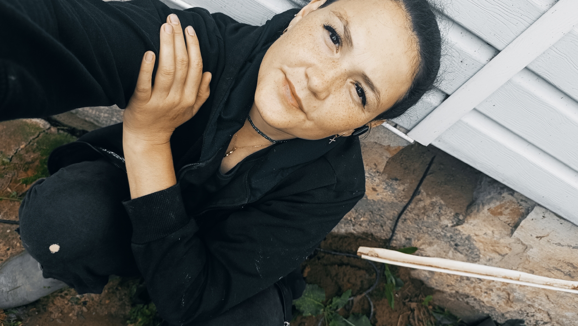 A high-angle, medium shot captures a person kneeling outdoors, against a backdrop of a white vinyl-sided structure and rocky ground. The person, with dark hair pulled back, wears a black zip-up hoodie and dark pants, with grey boots visible. They are positioned in the lower left of the frame, with their face turned towards the camera. The individual has fair skin with freckles and wears light makeup, including prominent eyelashes. They are adorned with several necklaces, including a dark beaded one and a delicate gold chain with a pendant, as well as small cross-shaped earrings. Their left hand rests on their chest, and their right arm is bent, with the hand near their face. The environment appears to be a backyard or undeveloped area, with scattered stones and some green foliage on the ground. The lighting suggests it is daytime, with a slightly overcast or diffused light source. The image does not contain any specific indicators of the city of Karsha, Russia.
