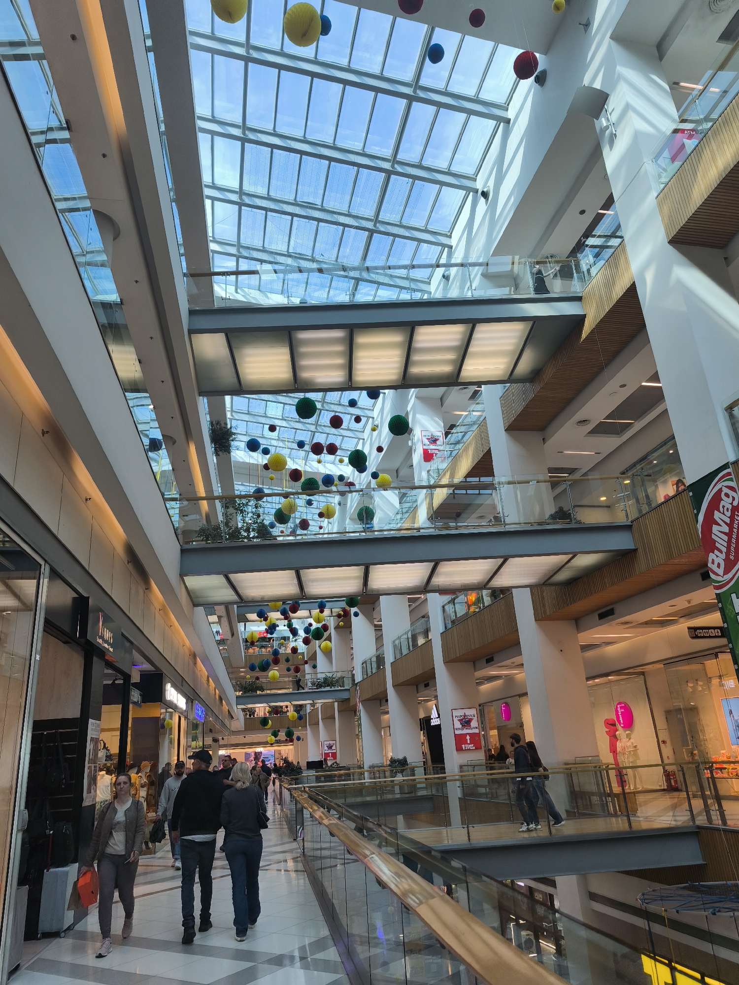 The image shows the interior of a modern shopping mall, likely in Varna, Bulgaria, based on the "BulMag Supermarket" sign visible. The mall has multiple levels connected by walkways with glass railings, and a large glass ceiling allows natural light to flood the space. Suspended from the ceiling are numerous colorful decorative spheres.
Several people are present. On the lower level, a group of shoppers walks along a corridor with retail stores on either side. Some are in groups, while others are alone. On an upper level walkway, a person stands near the railing. The mall appears well-lit and clean, with a contemporary architectural design featuring a mix of glass, steel, and wood elements.