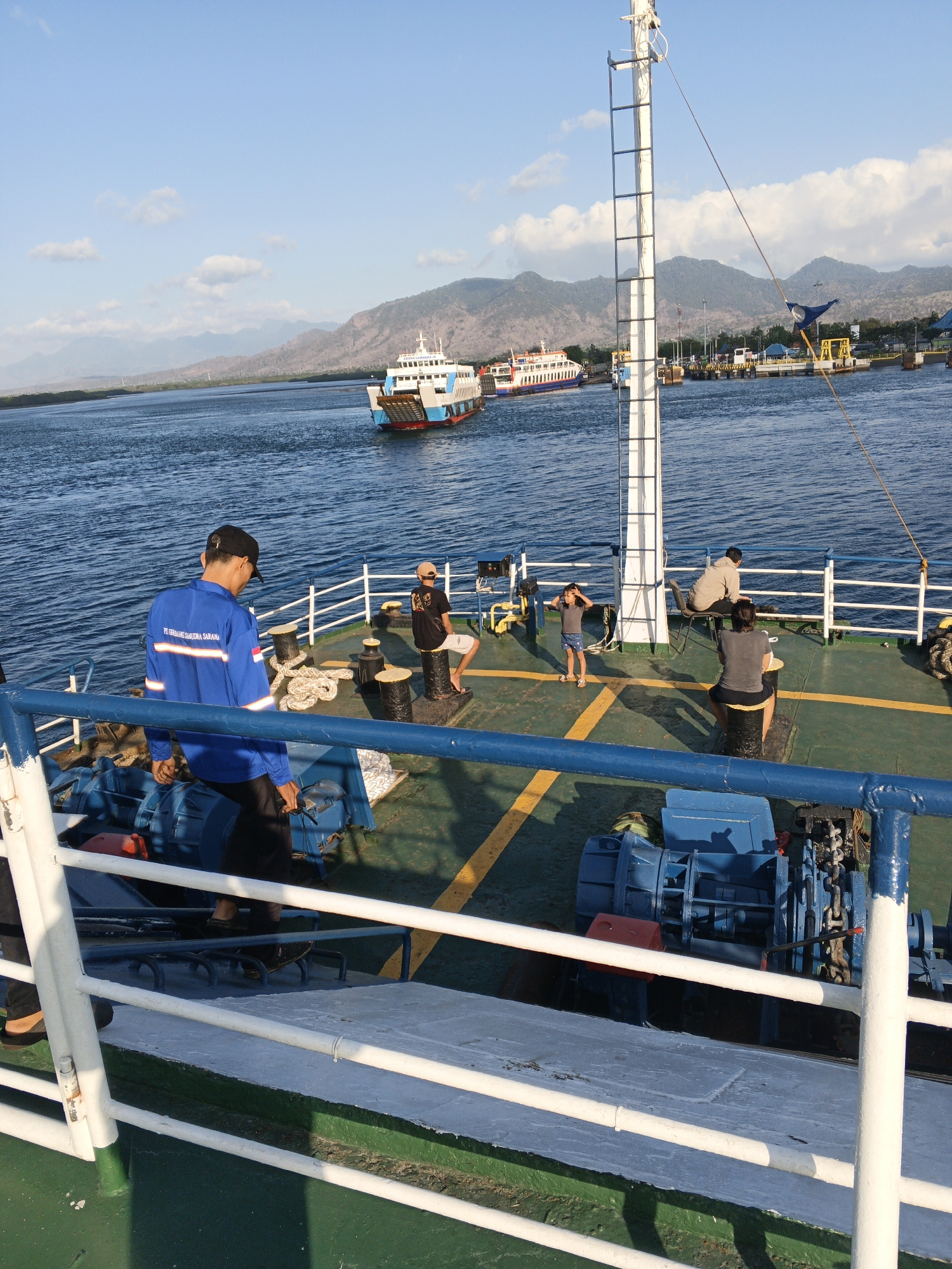 The image captures a scene on the deck of a ferry near the coast of Melaya, Indonesia. The sky is blue with scattered clouds, and a range of mountains can be seen in the background. The sea is calm and blue, with several other ferries visible in the distance, suggesting a busy port or shipping route.
On the deck of the ferry where the photo is taken, there are several people. In the foreground, a person wearing a bright blue uniform with reflective stripes and a black cap is looking down. To their right, a person in a black t-shirt and white shorts is seated. Further back, a child in a grey t-shirt and blue shorts stands with their arms raised near their head. Another person in a light-colored hoodie is seated on a chair. A person in a dark t-shirt and dark shorts is also present.
Objects on the deck include large black mooring bollards, coiled ropes, and a blue winch mechanism with a red container attached. The deck itself is painted green with yellow lines marking pathways. White railings with blue metal supports line the edge of the deck. The overall impression is a moment of transit on a ferry, with passengers and crew members interacting within the operational environment of the vessel.
