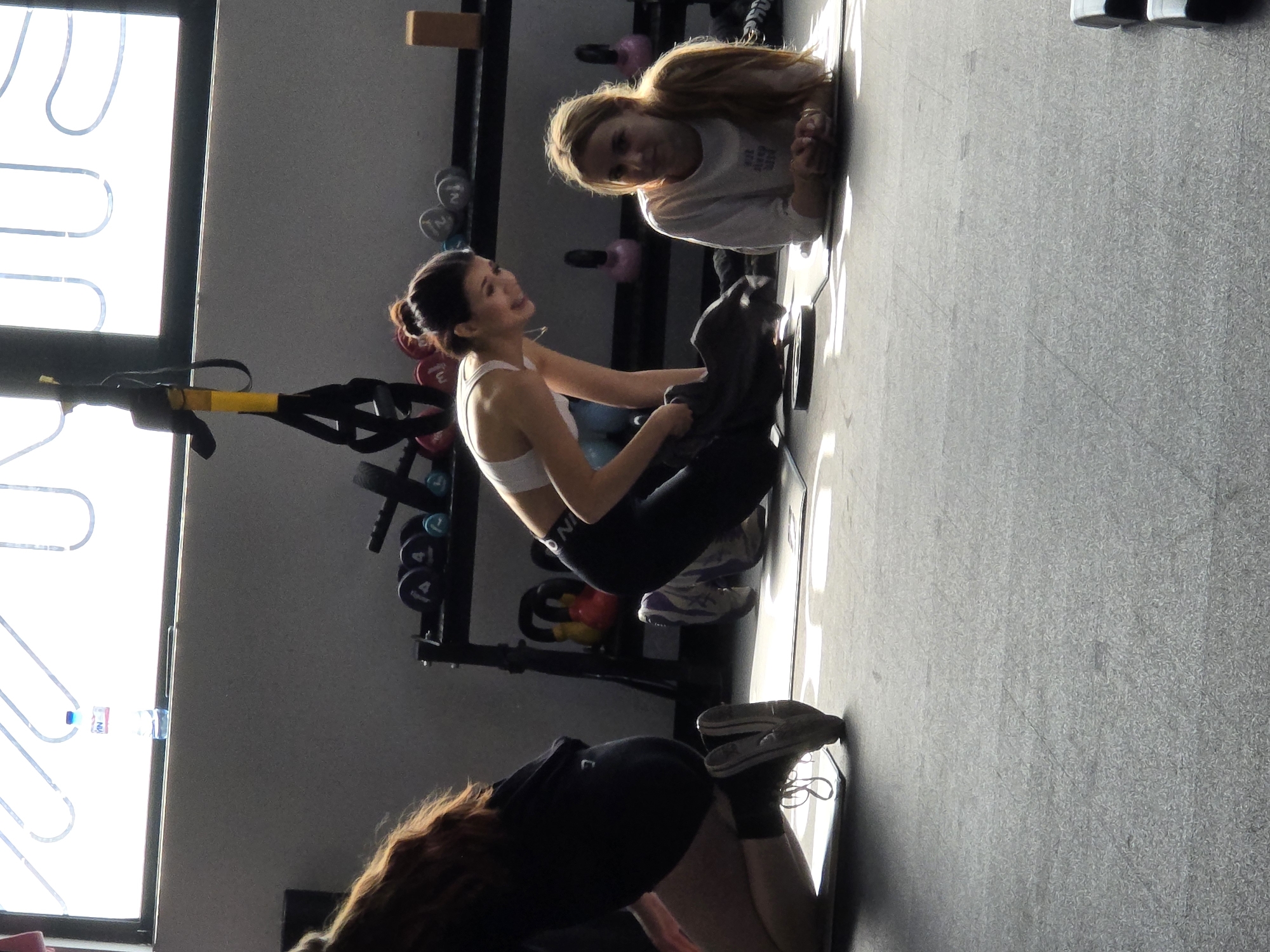 Three women are engaged in a fitness session inside a well-lit gym or studio. The space features grey walls and a dark grey, possibly rubberized, floor, suggesting a dedicated workout area. Bright natural light streams in from a large window on the left, which has white S-shaped patterns on its glass and a small bottle labeled "VN" on the sill.
In the foreground, a woman with long reddish-brown hair is seen from behind, kneeling on a mat, wearing a dark t-shirt and shorts. In the midground, a woman with dark hair tied in a bun is seated on a yoga mat, wearing a white sports bra (partially visible "NIKE" logo) and dark leggings. She appears to be performing an abdominal exercise or stretch, with her knees bent and feet on the floor. She is smiling broadly, looking towards the blonde woman or slightly off-camera. Behind her, a black fitness rack holds various dumbbells (some marked "2"kg and "4"kg) and colorful kettlebells (red and blue/pink). A yellow-handled TRX suspension trainer hangs from the rack.
Further back, on another mat, a blonde-haired woman is lying prone, propped up on her forearms, looking directly at the camera with a slight smile. She is wearing a light-colored long-sleeve top with faint text that seems to say "LIVE AND BE FREE". A wooden block is visible on top of the fitness rack behind her. The atmosphere appears positive and relaxed. The strong natural light indicates it is daytime. While the specific location of Plovdiv, Bulgaria, cannot be discerned from the image itself, the scene depicts a modern fitness environment.