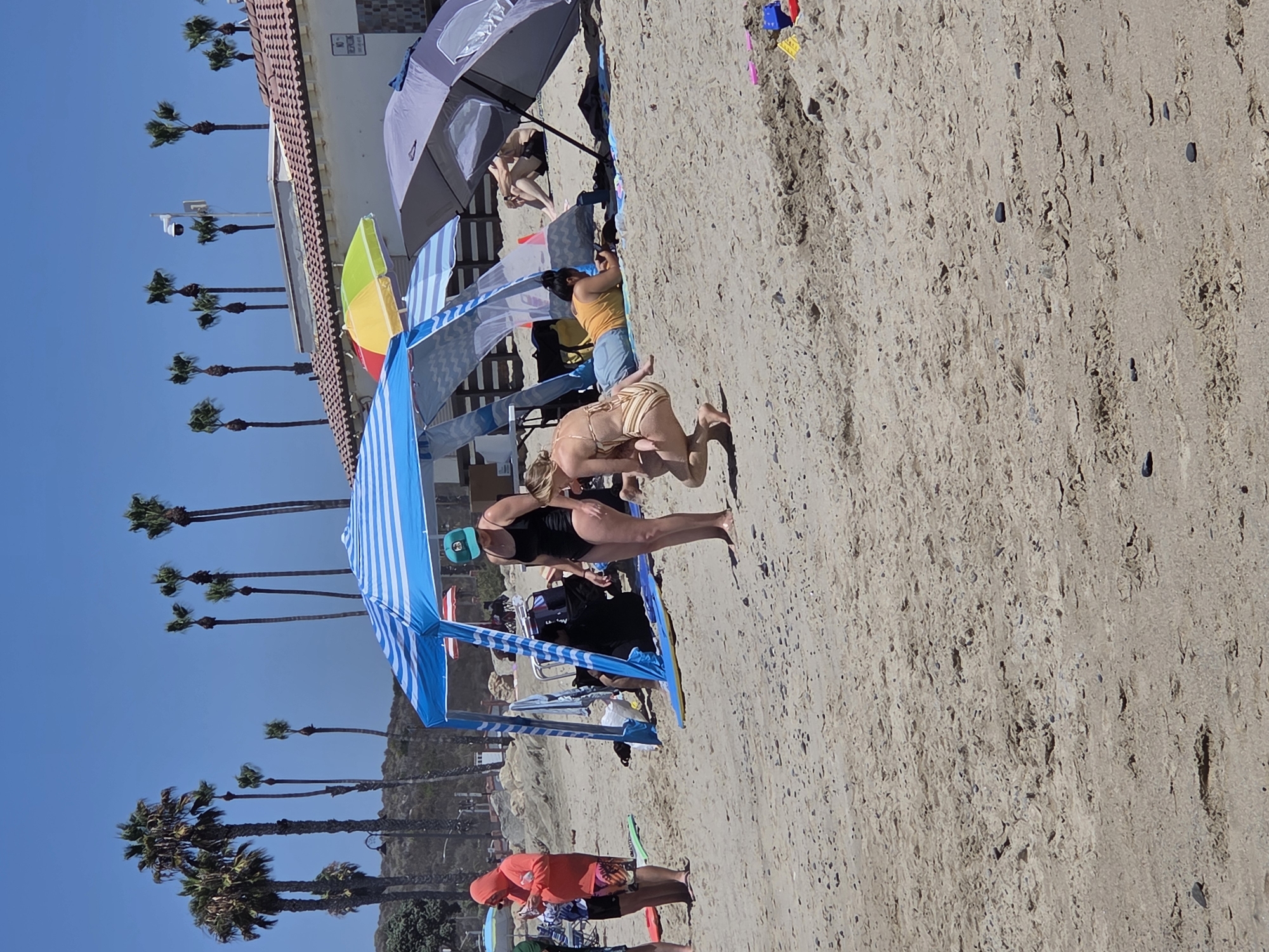The image depicts a sunny day at a beach in San Clemente, California, United States of America. Several people are present, some lounging under beach umbrellas and canopies, others standing. In the foreground, two women are interacting. One woman in a striped bikini is crouching down, appearing to hold or tend to a younger child, also in a bikini. Beside them, a woman in a black swimsuit and a green baseball cap stands facing away from the camera.
Further back, a man in swim trunks is seated under a large gray umbrella. To the left, a person in a bright orange hooded jacket and swim trunks stands holding a phone. The beach is sandy with scattered small stones. Palm trees are visible in the background, along with a building with a tiled roof and a security camera. Beach equipment, including umbrellas and a blue and white striped canopy, are set up on the sand. A few colorful toy building blocks are visible in the lower right corner. The sky is clear and blue.