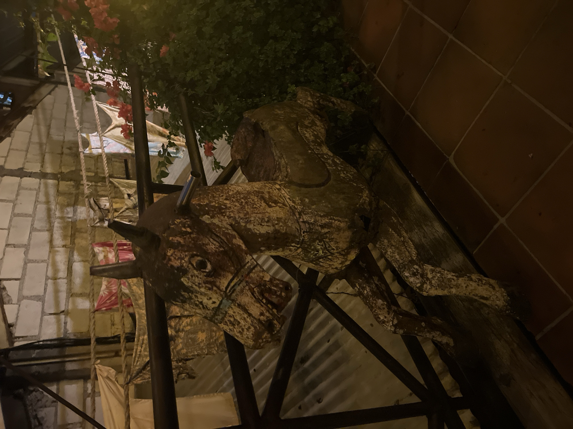 An old, weathered rocking horse sits on a wooden beam. The horse is made of wood and appears to be quite old, with peeling paint and a worn appearance. It is positioned on a balcony or patio with a tiled floor and a railing. In the background, there is a brick wall and some hanging fabric. A lush green plant with red flowers is visible to the right of the horse. The scene is lit by artificial lighting, creating a warm, somewhat dim atmosphere. The location is identified as Kuta, Indonesia. There are no people or animals visible in the image.