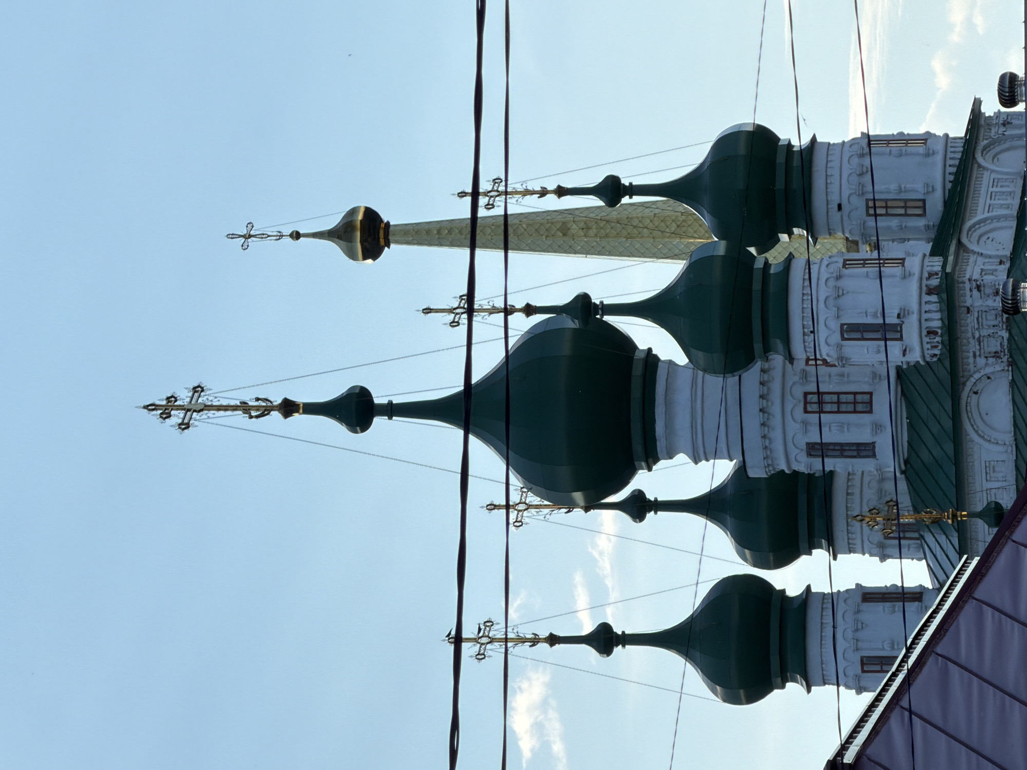 The image shows the gilded crosses atop several domed towers of the Holy Trinity Cathedral in Solikamsk, Russia. The cathedral, constructed in the 17th century, is a significant example of Russian Baroque architecture. The dominant colors are the light blue of the sky and the dark green of the domes, contrasted with the white of the cathedral's walls. The sun is high in the sky, suggesting it is daytime, and there are some wispy clouds present. The perspective is looking upwards at the cathedral's spires, with electrical wires crisscrossing the frame. The overall impression is one of grandeur and historical significance, with the bright sunlight highlighting the golden crosses against the sky. The scene is tranquil, with no visible people or activity other than the birds perched on the crosses. The architectural details of the cathedral, such as the arched windows and ornate carvings, are visible on the white walls.