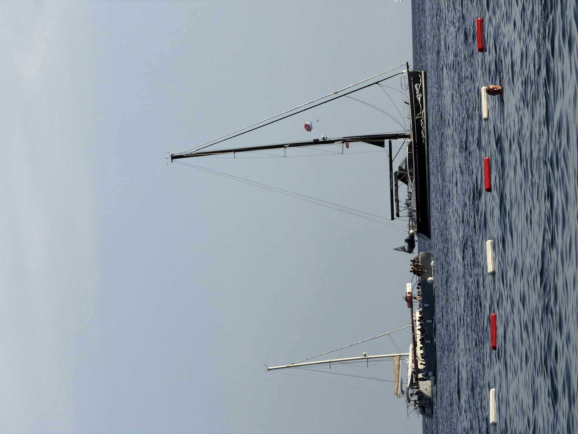 This image depicts a serene seascape off the coast of Budva, Montenegro. The foreground is dominated by the gentle ripples of the Adriatic Sea, punctuated by three floating buoys: two red and one white, arranged in a horizontal line. In the middle ground, a larger, dark-hulled sailboat with a prominent black mast stands to the right, its sails furled. A parasailor, a colorful red and white canopy, is visible in the sky behind the sailboat, with two figures suspended beneath it. To the left of the sailboat, a long, gray catamaran-style boat is visible, also at anchor. This vessel is crowded with numerous people, suggesting it is a tour or excursion boat. The background is a clear, pale blue sky. The overall scene suggests a leisurely summer day with various water activities taking place.