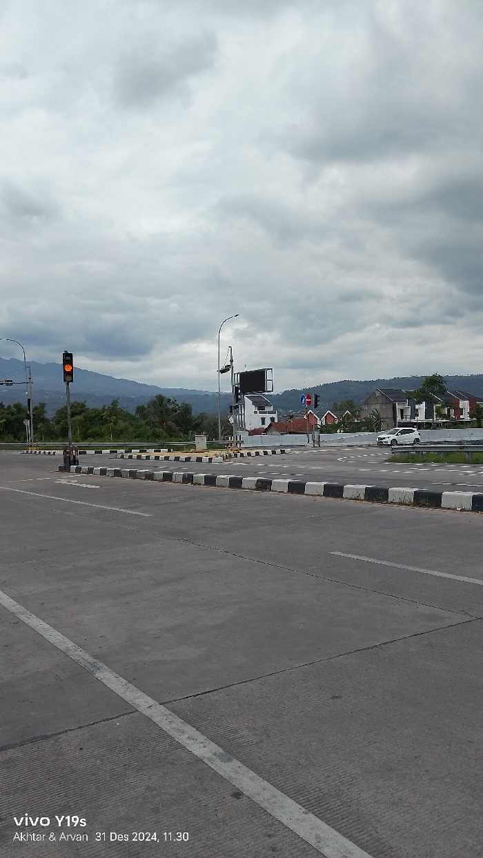 The image presents a wide outdoor view of a modern road intersection or interchange, likely in Sumedang, Indonesia, on an overcast day. In the foreground, a multi-lane concrete road stretches forward, marked with white lines defining lanes and an approaching intersection. The median strips and traffic islands within the intersection are clearly delineated by black and white painted curbs.
A prominent traffic light pole on the left displays an illuminated amber (yellow) light, indicating a cautionary state. Further into the intersection, other traffic signals and a blue circular "no entry" sign are visible. To the right of the intersection, a white car is seen, possibly navigating or waiting. On the right side, beyond the road, several modern, light-colored buildings with dark roofs are nestled amidst green foliage. In the mid-ground, a large, dark rectangular sign or billboard is mounted on a pole.
The backdrop consists of a range of distant, green-hued mountains under an entirely overcast sky filled with diffused grey clouds, suggesting a muted daylight. No people are visible, lending the scene a quiet, somewhat empty atmosphere. The bottom left corner of the image contains a watermark: "vivo Y19s Akhtar & Arvan 31 Des 2024, 11.30", providing details about the device, photographers, and the time the photograph was taken.