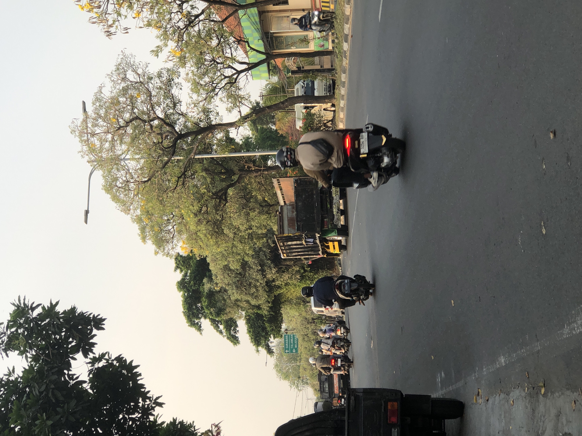 A street scene in Kludan, Indonesia, shows several motorbikes and a truck on an asphalt road. Two people are riding on a motorbike in the foreground, facing away from the camera. They are wearing helmets and casual clothing. The motorbike has a license plate with the number "3254 BB5". To the left, another motorbike is visible with two passengers. Further down the road, a truck is seen with cargo. Trees with green leaves and some yellow flowers line the sides of the road. A street light is also visible. The sky is overcast with a pale yellow hue. A signpost with directional arrows pointing to "SURABAYA", "TULANGAN", and "MOJOSARI" is visible on the left side of the road. The environment appears to be a suburban or semi-urban area with moderate traffic.