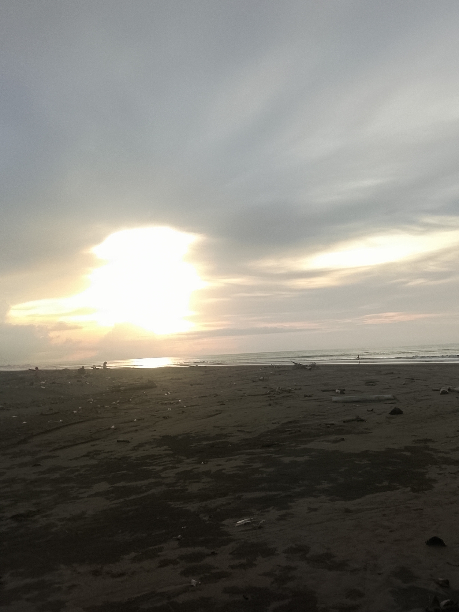 This is a full shot of a beach scene at what appears to be dusk or dawn. The sun is bright, partially obscured by clouds, and casts a glow on the ocean. The sky is overcast with soft light filtering through. The beach is dark sand, littered with driftwood, debris, and some plastic. There are a few small figures of people walking on the beach in the distance. The waves are small and gentle. The scene depicts a tranquil, natural environment with evidence of human impact through the scattered debris.