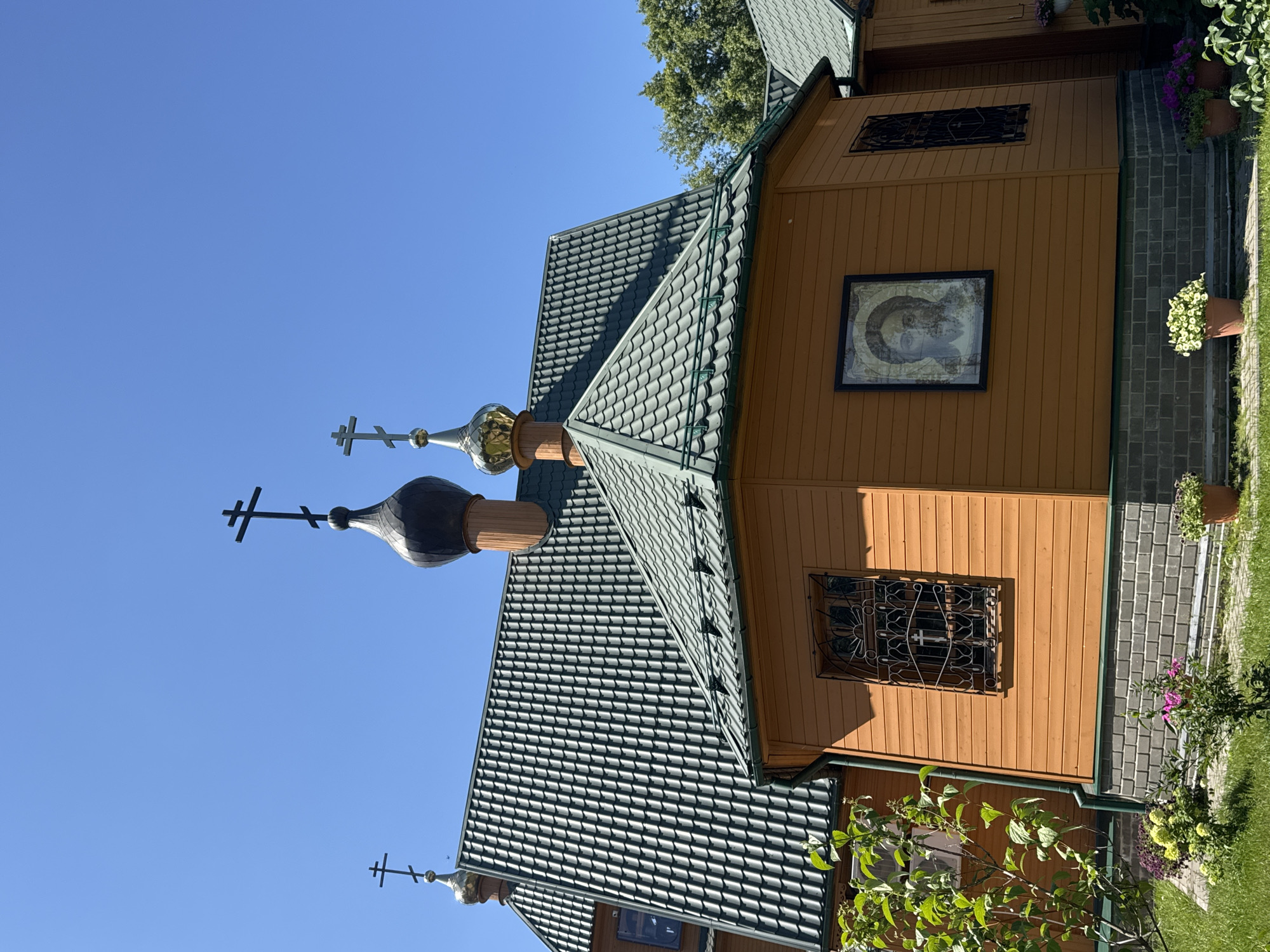 This is an exterior, eye-level, slightly low-angle, medium shot of a wooden church with a green tiled roof under a clear blue sky. The church has two onion domes topped with Orthodox crosses. One dome is black, and the other is gold. The wooden walls are orange-brown. There are two windows visible on the main section of the church. The window on the left is partially obscured by greenery and has a white frame. The window on the right is larger and features an ornate, black metal grate. Above the grate, a framed icon depicting a portrait of Jesus Christ hangs on the wall. The church is situated on a foundation of grey brick. In the foreground, there is green grass, a paved walkway, and several potted plants with colorful flowers. The church is located in Nizhny Novgorod, Russia.