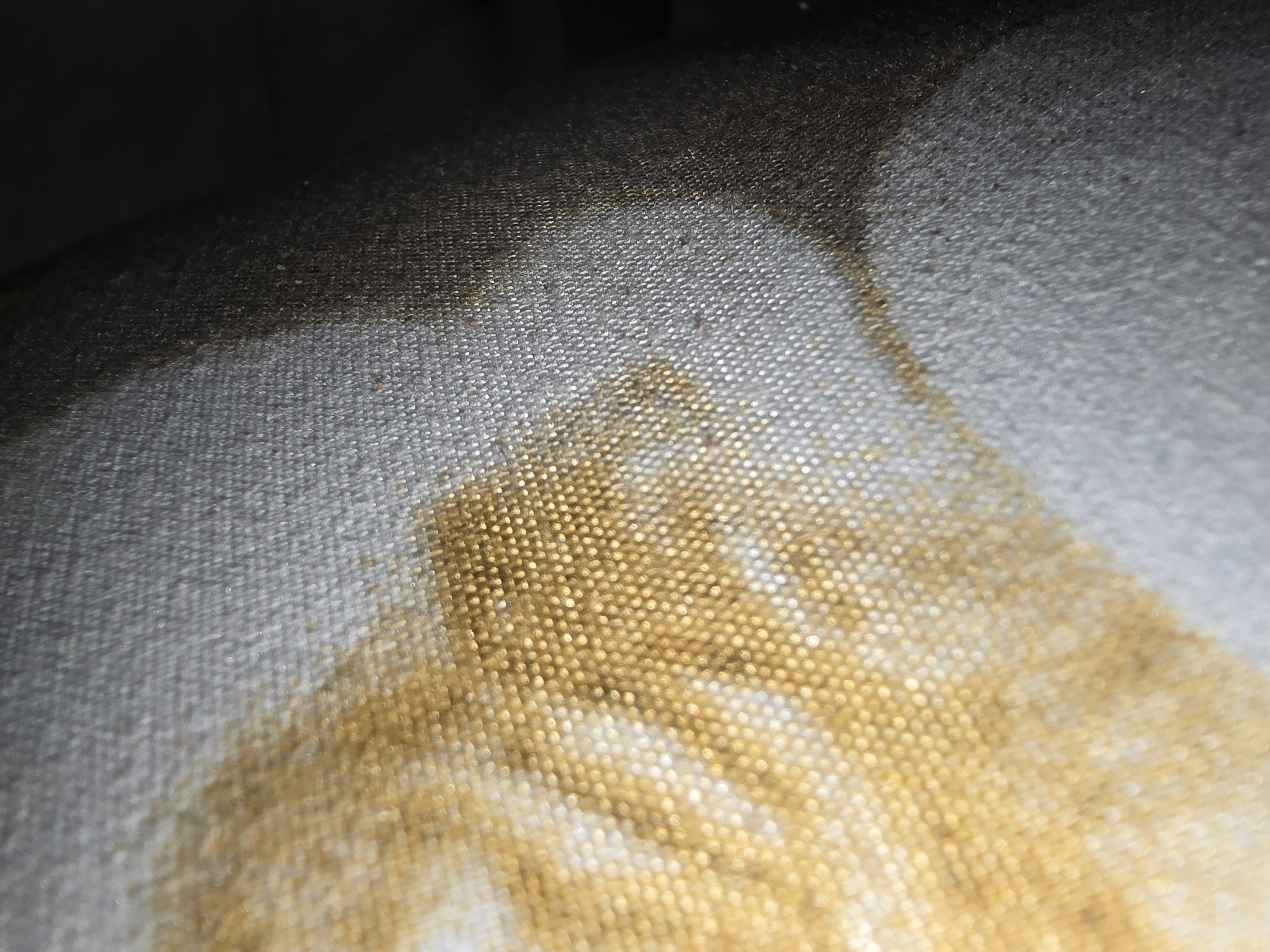 The image displays a close-up, low-angle shot of a coarse, textured fabric, possibly upholstery or a canvas. The fabric is predominantly gray with distinct patches of a yellowish-gold stain, concentrated on the left side and fading towards the right. The texture of the fabric is visible, with small, evenly spaced woven threads creating a grid-like pattern. Some areas of the fabric appear darker, transitioning into a deep shadow on the far right, suggesting an absence of light or an object casting a shadow. There are no discernible people, animals, or specific landmarks visible in the image. The scene is devoid of any context that would place it in Ulan-Ude, Russia, or any other specific geographical location. The image is purely a textural and color study of the stained fabric.