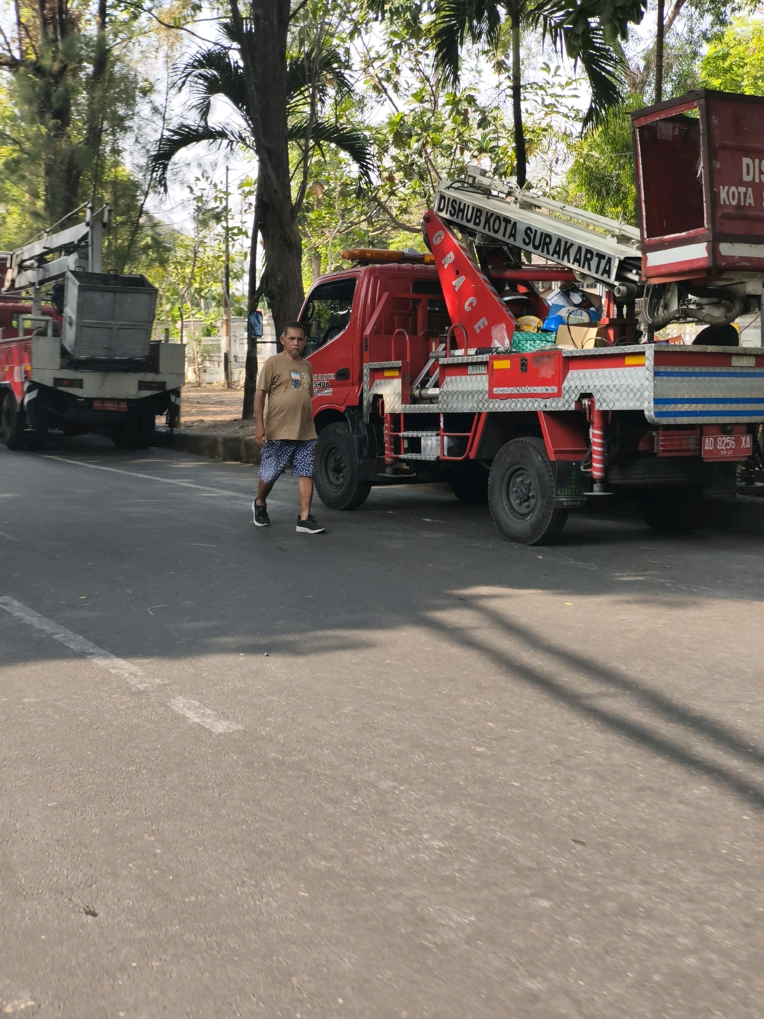 A man walks along a paved road in Surakarta, Indonesia, in front of two red trucks with crane attachments. The truck on the right has a license plate reading "AD 8256 XA." The truck on the left has a grey metal bin attached to its crane. The man, wearing a light brown t-shirt and patterned shorts, walks from right to left, facing forward with his arms slightly bent. Trees and foliage line the right side of the road. The sky is visible through the trees.