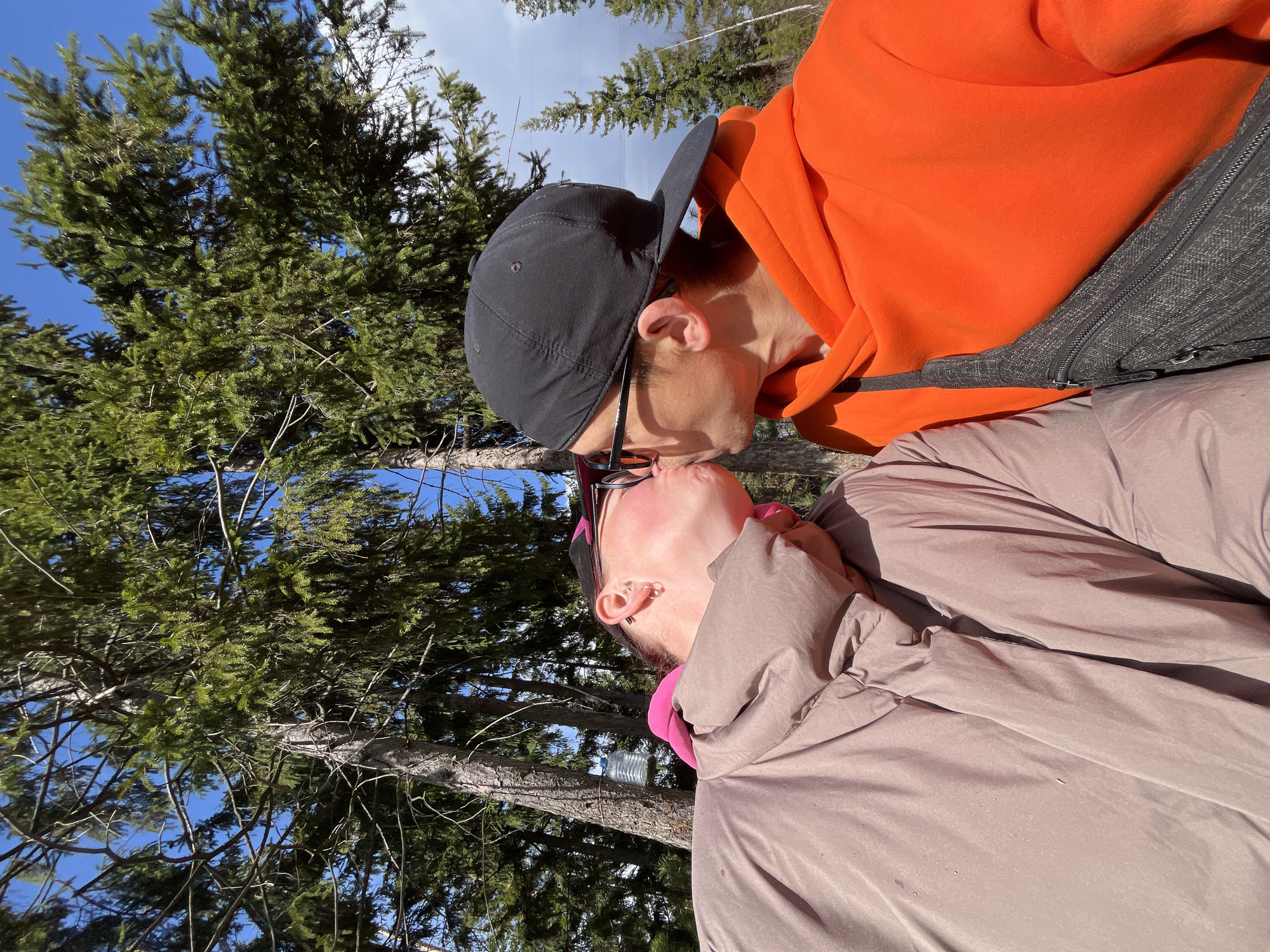 This outdoor image, taken in Chaikovsky, Russia, captures two individuals kissing amidst a backdrop of tall pine trees under a clear blue sky. The bright sunlight suggests it is daytime, likely midday or afternoon.
One person, positioned in the upper portion of the frame, wears a vibrant orange hooded sweatshirt and a black baseball cap worn backward. A grey fanny pack or sling bag is visible across their chest. They are leaning in to kiss the other individual.
The second person, in the lower part of the frame, wears a light brown or taupe puffy jacket over a pink inner layer. They appear to have glasses or sunglasses pushed up onto their head and multiple ear piercings. Their head is tilted upwards in the embrace.
The interaction conveys a sense of affection and intimacy within a natural, serene environment. In the background, among the evergreen trees, a clear plastic water bottle can be seen hanging from a branch. No discernible text is visible in the image.