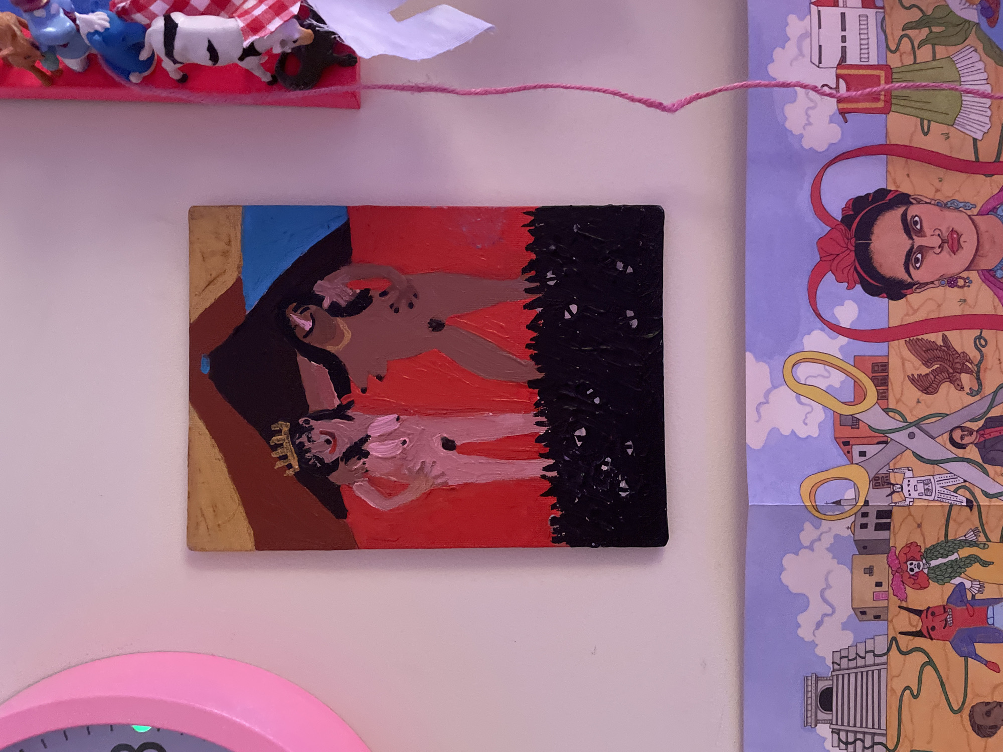 The image displays a collection of art and decorative items arranged on a light-colored wall, likely in an indoor setting. On the left, a large, pink, circular object, possibly a clock face, is partially visible. To its right, a small, rectangular painting depicts two stylized, nude figures against a red background. The figures are outlined in black, with one wearing a gold crown. A dark, textured area representing foliage or ground is at the bottom of the painting. To the right of the painting, a vertical pink border holds several small, toy-like figures, some of which are brightly colored. A thin, pink string hangs vertically in the center of the frame, originating from somewhere above the painting. Below the painting, a panoramic artwork is displayed, featuring various elements of a landscape with architectural structures and stylized figures, including a portrait of Frida Kahlo on the right. The overall scene appears to be a personal display of artistic taste. The image does not contain any identifiable elements that suggest a specific location within Vracar Urban Municipality, Serbia.