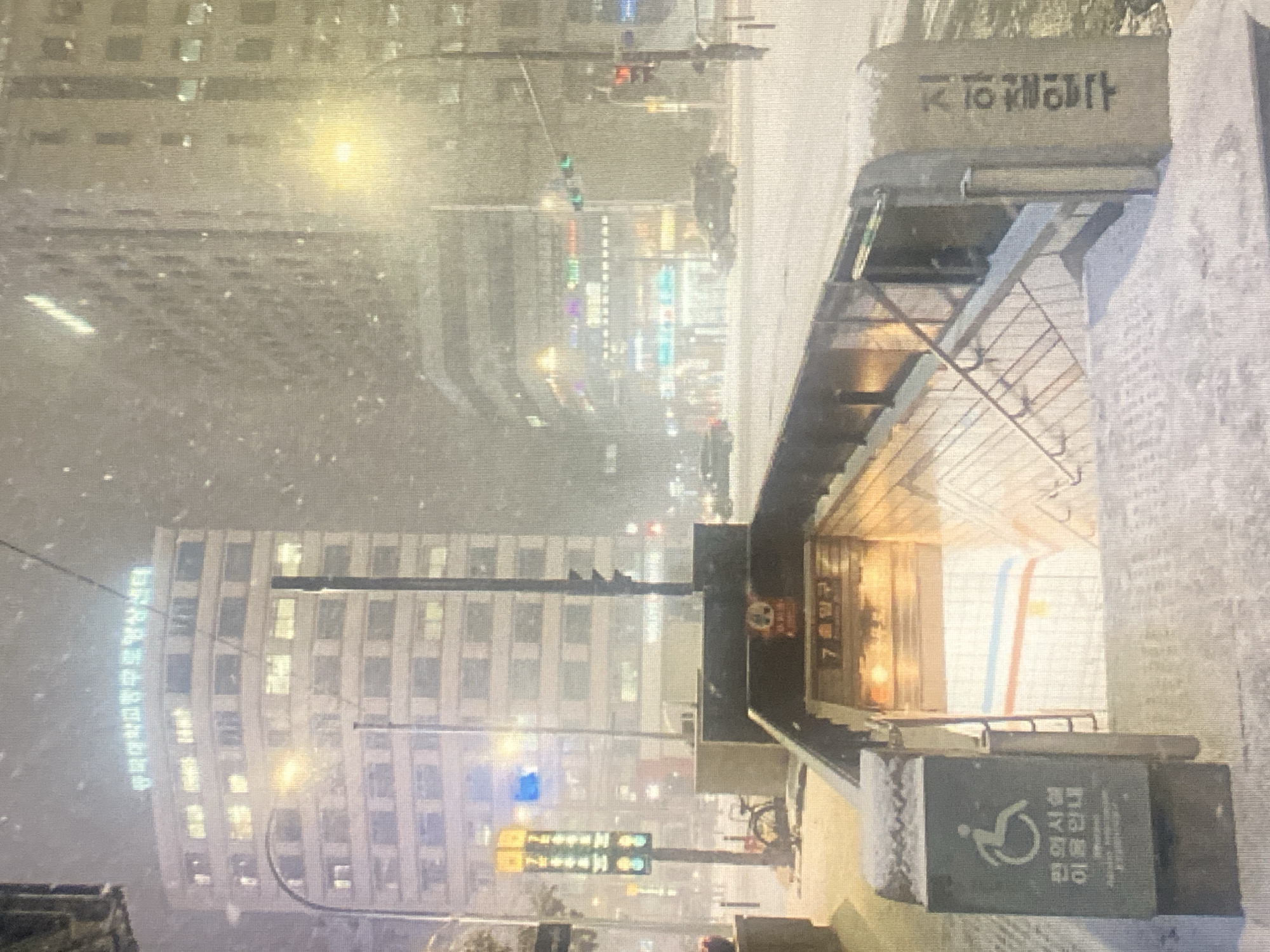 The image depicts a snowy urban scene at night, with heavy snowfall obscuring details. A multi-story building with illuminated windows is visible in the background, bearing Korean text in neon lights that reads "한국대학기록정보센터". In the foreground, a subway entrance is visible, with stairs leading down. A sign with a wheelchair icon and Korean text is attached to a structure next to the entrance, indicating accessibility information. Another sign with Korean text, "7호선출구", is visible above the subway entrance, indicating the subway line number. A traffic light with red and green signals is also present in the middle ground. Cars are driving on the snow-covered street. The overall environment suggests a cold, winter night in a city.