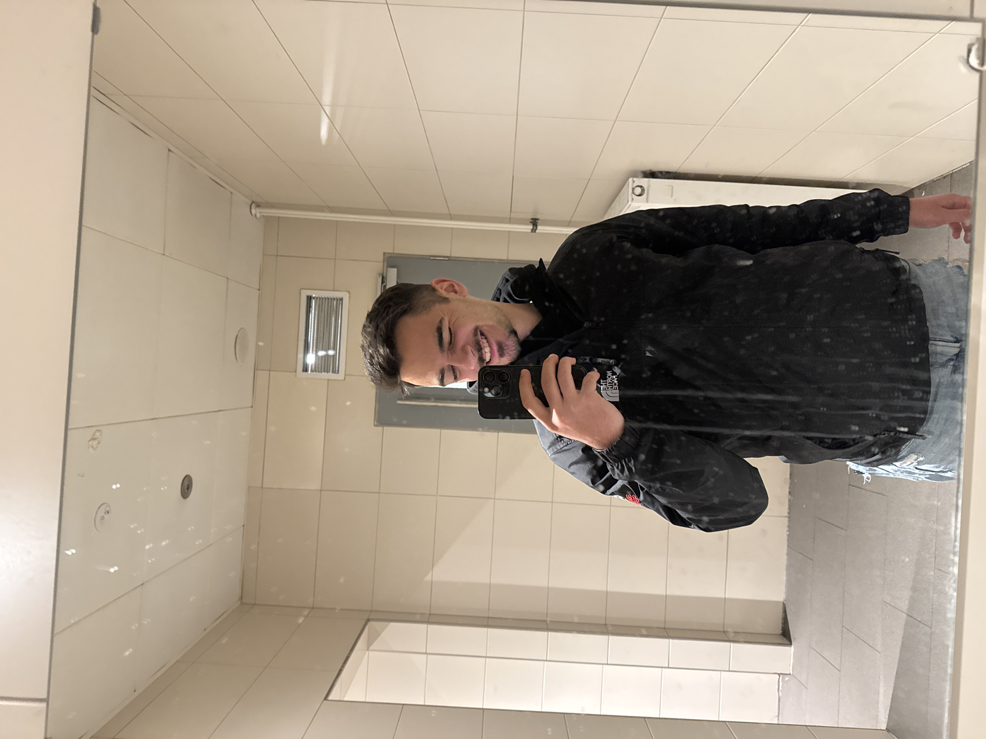 A young man with short dark hair and a light beard is captured in a mirror selfie, smiling broadly with visible teeth. He is wearing a dark, possibly black, jacket with white speckles, and blue jeans. He holds a black smartphone with multiple rear cameras in his left hand, taking the picture. The jacket features a mirrored "THE NORTH FACE" logo.
The setting is an indoor public restroom or similar utilitarian space, characterized by light-colored, rectangular tiled walls, a tiled ceiling, and a tiled floor. In the background reflection, a metallic ventilation grate is visible on the left wall, along with a white radiator or heater further back. A white toilet tank or urinal is partially visible behind the man's right shoulder. A grey door and other white wall fixtures can also be discerned.
The man's expression conveys happiness and cheerfulness. The time of day is not indicated, as the scene is artificially lit. The speckles on his jacket might suggest recent exposure to precipitation. The general aesthetic points to a public facility in Sofia, Bulgaria, as per the provided context. Faint text like "LIVE" and "0.5x" can be seen reflected on the phone's screen, indicating its camera mode.