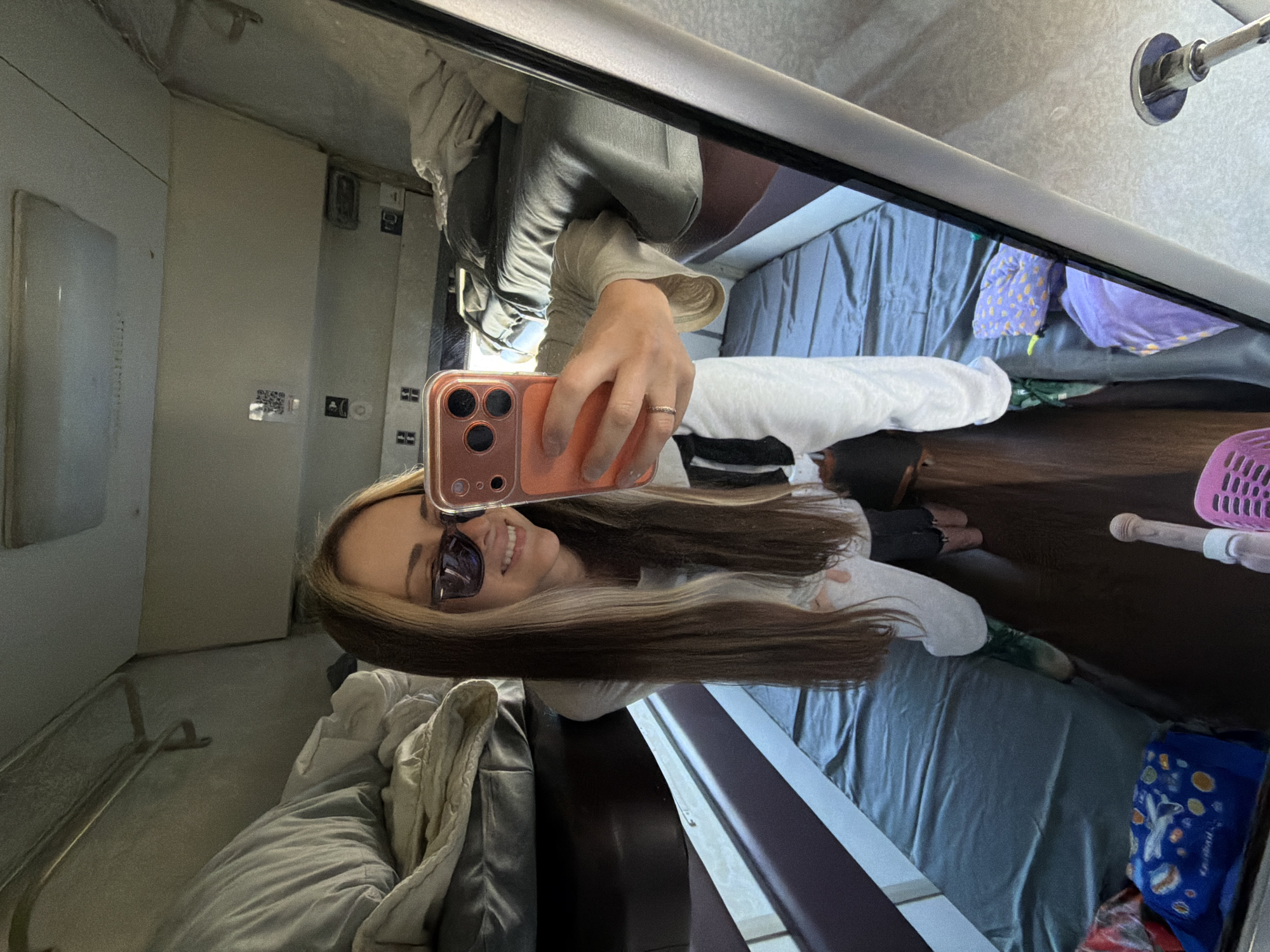 A woman is taking a selfie in a mirror inside what appears to be a train compartment. She is wearing sunglasses and holding a phone. The compartment has blue bedding, a wooden table, and personal items like a pink basket and a blue bag. The location cannot be determined as Lebyazh'e, Russia as there is no evidence to suggest this.