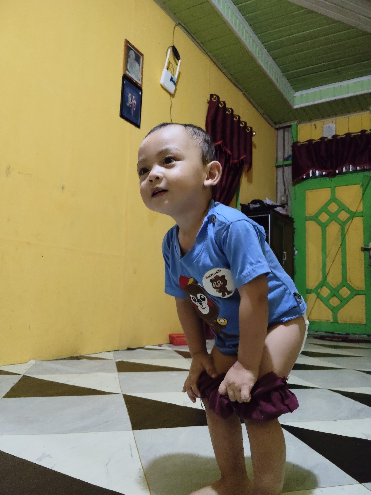 A young child is shown in a room with yellow walls and a green ceiling. The child is wearing a blue t-shirt with a cartoon image and is pulling down maroon pants. The floor is tiled with a black and white diamond pattern. In the background, there is a green door with yellow geometric designs and a dark cabinet. On the wall, two framed pictures hang. A white light fixture is also visible on the wall. The scene appears to be indoors, in a residential setting in Tanah Grogot, Indonesia.