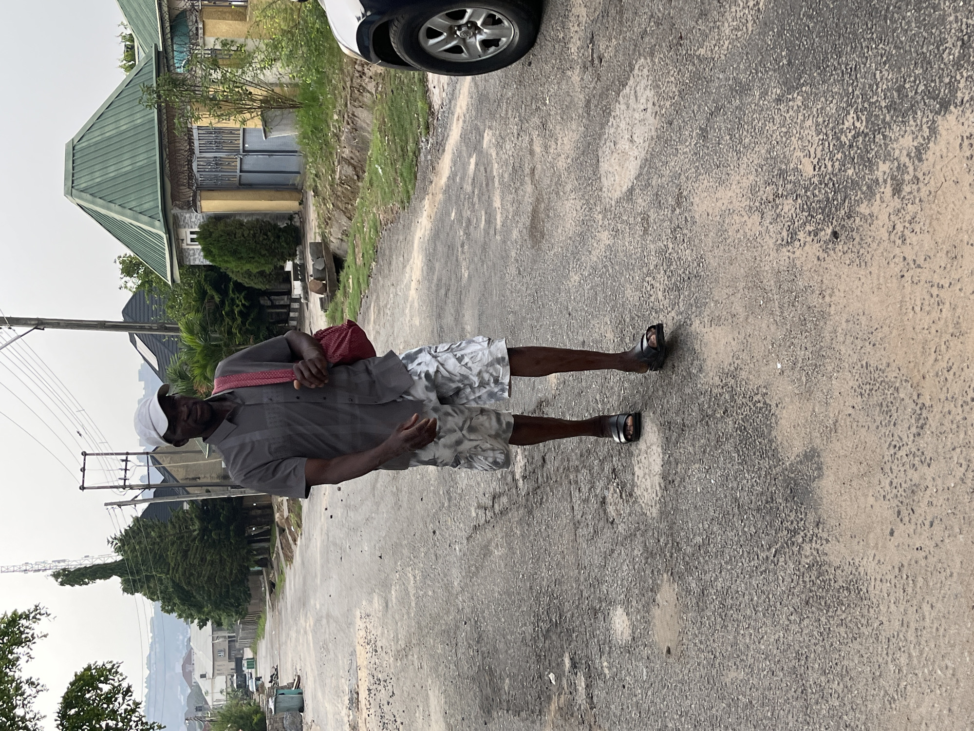 An elderly man is standing on an asphalt road in what appears to be a residential area of Abuja, Nigeria. He is wearing a gray short-sleeved shirt, patterned gray and white shorts, and sandals. He has a white cap on his head and a red bag over his shoulder. The road is cracked and shows signs of wear. In the background, there are houses with green roofs, trees, power lines, and a distant cityscape. A portion of a silver car is visible in the upper right corner of the image.