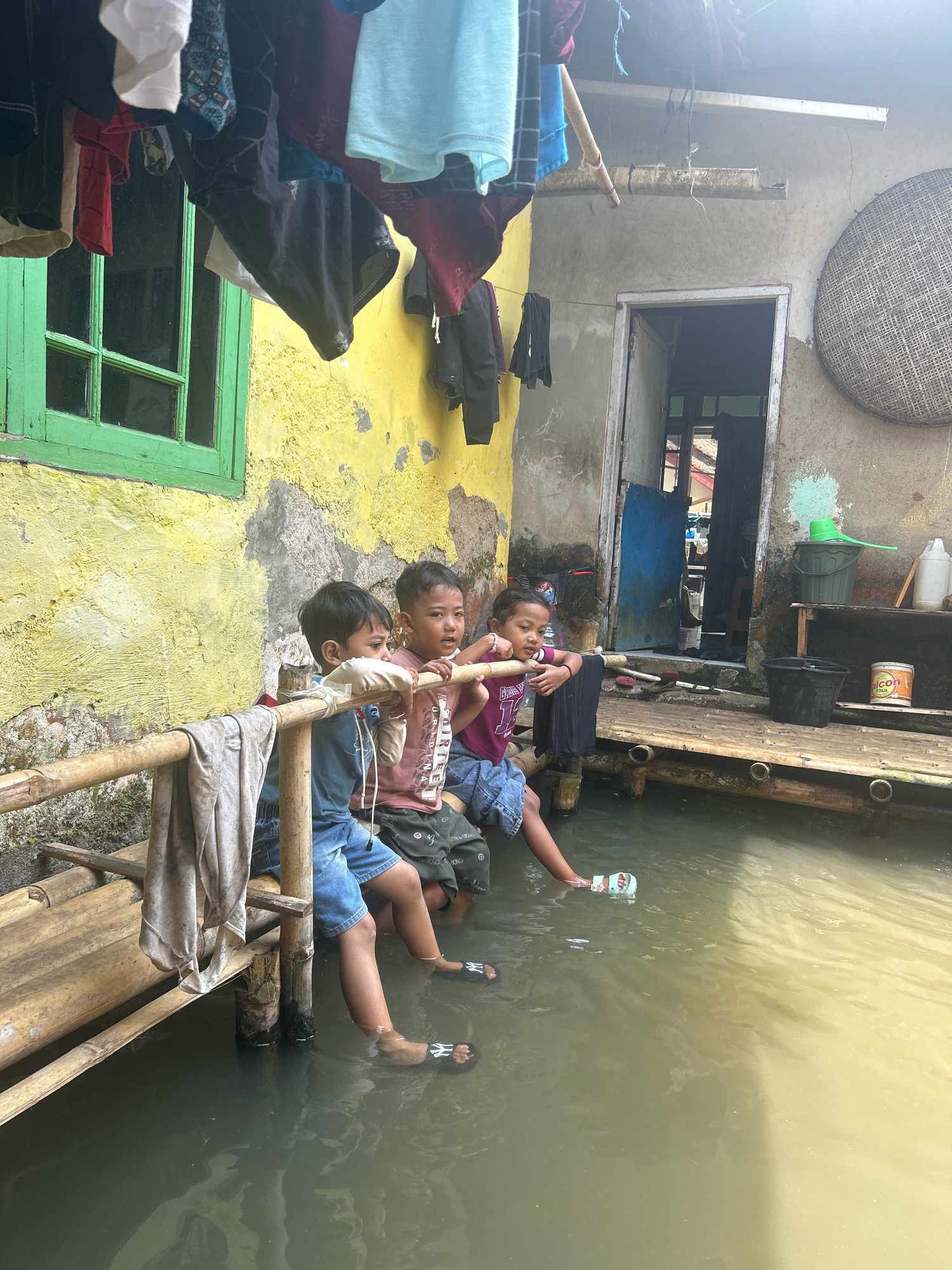Three young children sit on a bamboo platform that is partially submerged in murky water. The children are all facing to the right and appear to be looking into the water. The child on the left is wearing a blue t-shirt and denim shorts. The child in the middle is wearing a pink t-shirt and dark shorts. The child on the right is wearing a purple t-shirt and blue shorts. They are all wearing flip-flops. Behind them, a weathered yellow wall with a green window is visible, along with a doorway leading into a dark interior. Clothes are hanging on a line overhead. The scene depicts a home in Cihaurbeuti, Indonesia, likely affected by flooding, with the children finding a way to interact with the water that has inundated their living space.