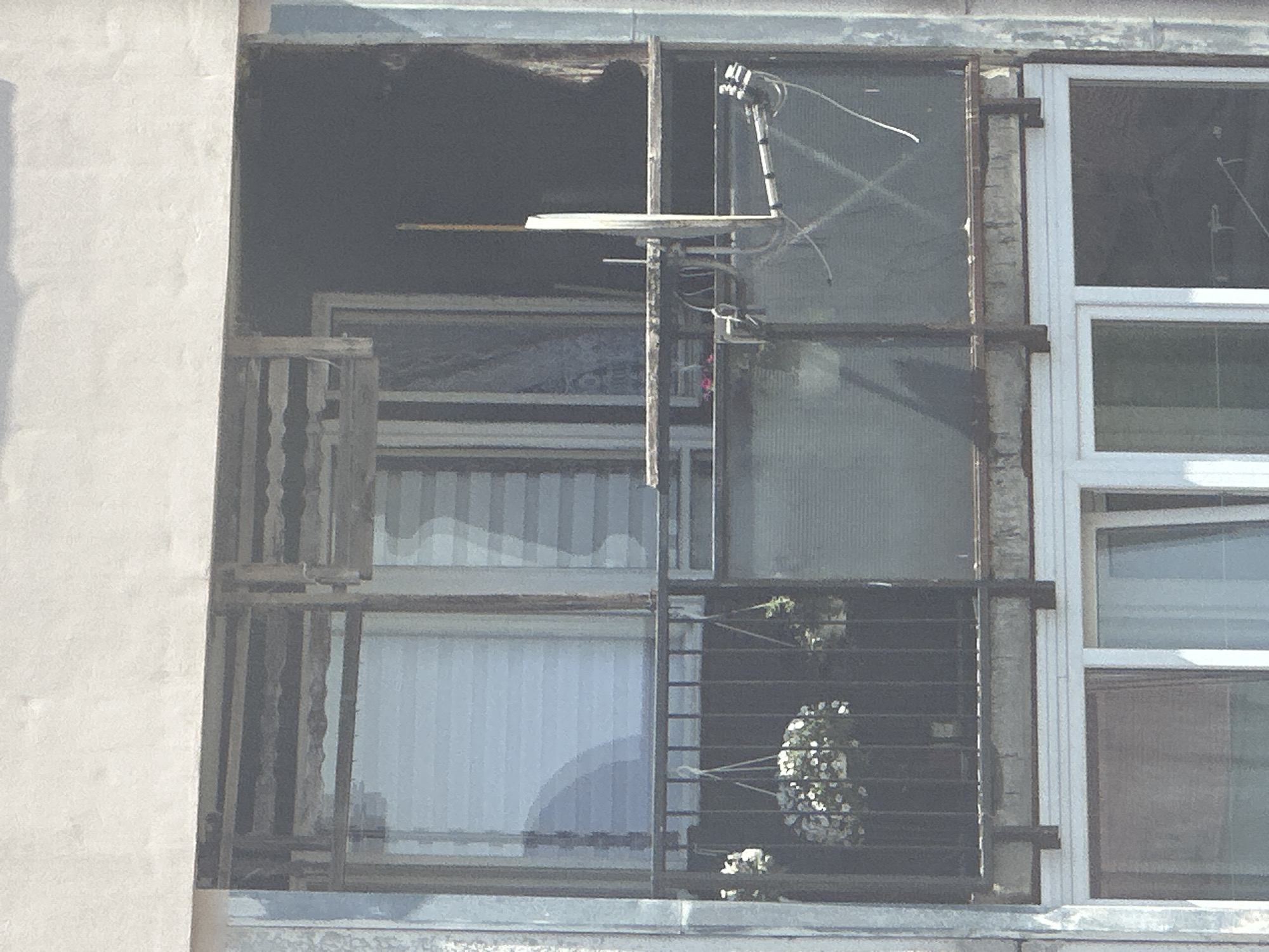 The image shows the facade of a multi-story building, likely residential, in the city of Uzhhorod, Ukraine. The focus is on an open balcony on an upper floor. The balcony is enclosed by a metal railing and has a floor that appears to be concrete. On the balcony, there are several hanging planters with white flowers. There is also a satellite dish mounted on a pole extending from the balcony structure. The windows behind the balcony appear to be modern, with some showing blinds. The overall impression is of an aging building with some signs of wear, but the balcony itself is decorated with plants. The weather appears to be sunny, casting shadows.