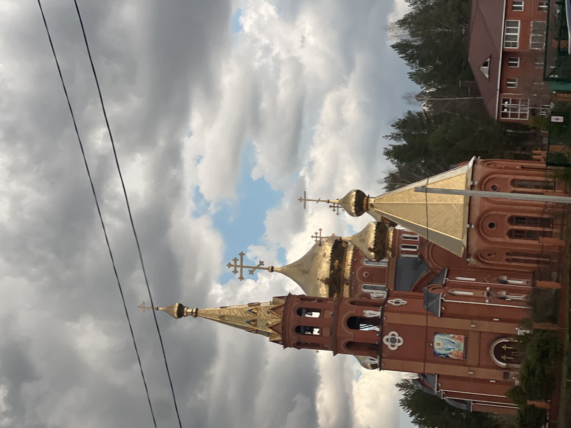 The image displays an Eastern Orthodox church with distinct Russian architectural characteristics, set against a partly cloudy sky. The church is constructed from red brick and features multiple golden onion domes of various sizes, each surmounted by a golden Orthodox cross. A prominent bell tower with arched openings and a golden spire stands on one side of the church.
The facade of the church includes arched windows, white decorative architectural elements, and a vibrant religious icon or fresco depicting holy figures, possibly the Virgin Mary and Christ, embedded on a side wall. The main entrance area is partially visible, showing an arch and additional small crosses.
In the background, a dense cluster of dark green coniferous trees, likely pines, stands behind the church. To the right and further back, a multi-story building made of brown-red brick with numerous windows is visible. The sky is largely covered by gray clouds, but patches of bright blue sky can be seen breaking through. Two dark utility lines diagonally cross the upper left portion of the frame. The scene is illuminated by natural daylight, with the golden domes reflecting sunlight, suggesting it is daytime. The architecture aligns with the context of Chaikovsky, Russia. There is no legible text visible in the image.