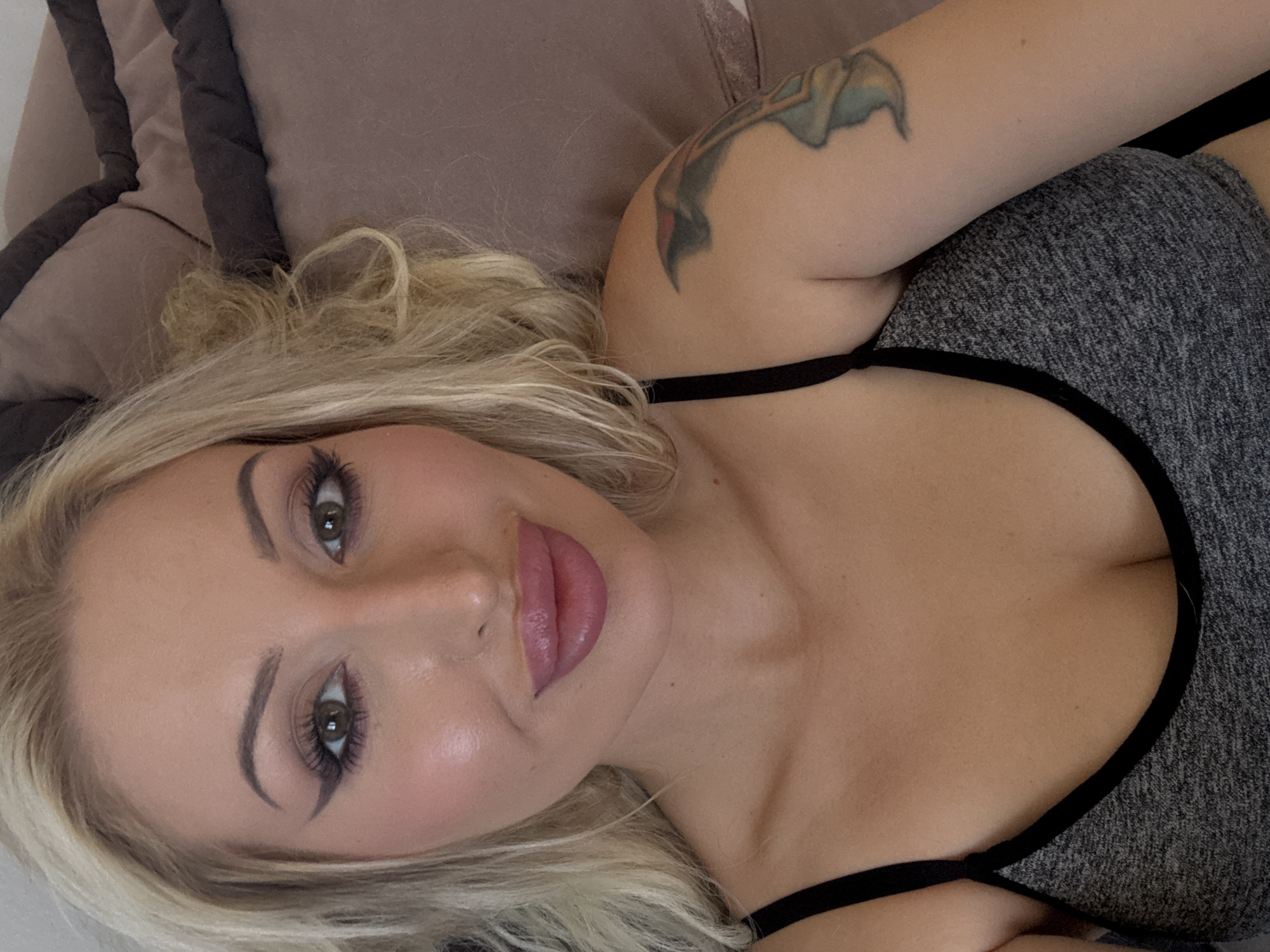 A close-up, medium shot shows a blonde woman lying on a sofa. Her face is angled towards the camera, and she is looking directly at it. She is wearing a grey sports bra with black trim. Her right arm is visible, and she has a tattoo on her upper arm. The background is a blurred brown sofa.