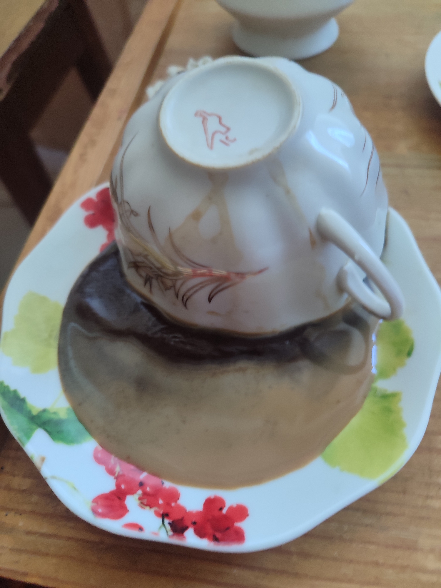 A white teacup, decorated with delicate brown and red floral patterns, is overturned on its saucer. The saucer is also white and adorned with similar floral designs, featuring red berries and green leaves. A dark liquid, likely coffee or tea, has spilled from the cup and pooled on the saucer, creating a glossy, reflective surface. The cup itself has a small red emblem on its underside. The scene is set on a wooden surface, possibly a table, with hints of other objects in the background, including what appears to be another white porcelain dish and a portion of a wooden structure. The lighting suggests it is daytime, and the overall impression is one of a domestic or cafe setting. No specific time of day, weather, or emotional cues are discernible from the image. The only visible text is the red emblem on the teacup.