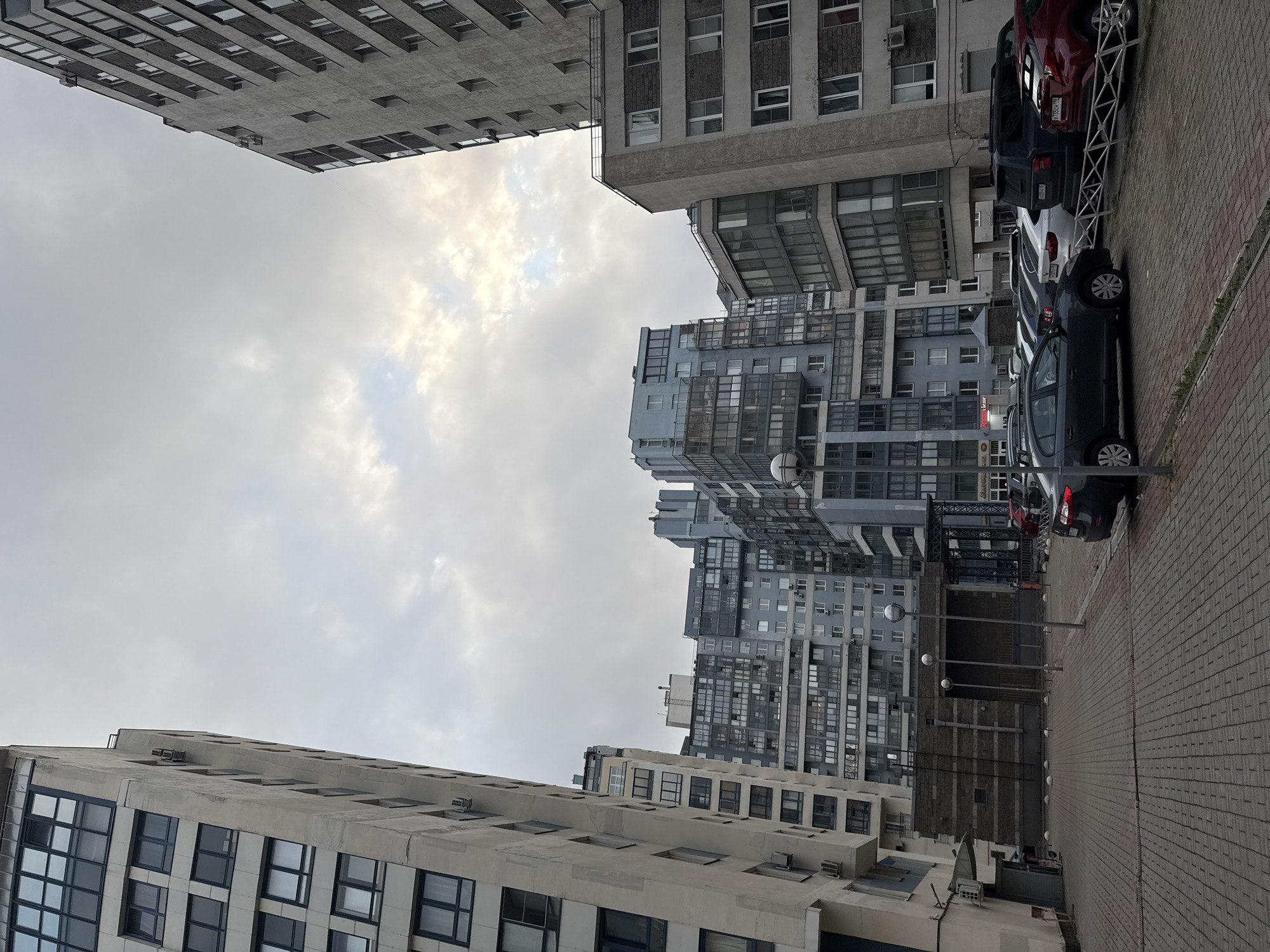 The image shows a multi-story residential buildings and a parking lot in an urban environment. The buildings are constructed with a mix of concrete, glass, and possibly brick, with some featuring balconies and air conditioning units. Several cars are parked in the lot, including a dark-colored sedan, a white SUV, and a red SUV. The sky is overcast, suggesting cloudy weather. The scene appears to be a typical residential area. The street is paved with interlocking concrete blocks. There are streetlights present in the parking lot. The architecture suggests a modern, possibly post-Soviet era development, consistent with many areas in Saint Petersburg, Russia.