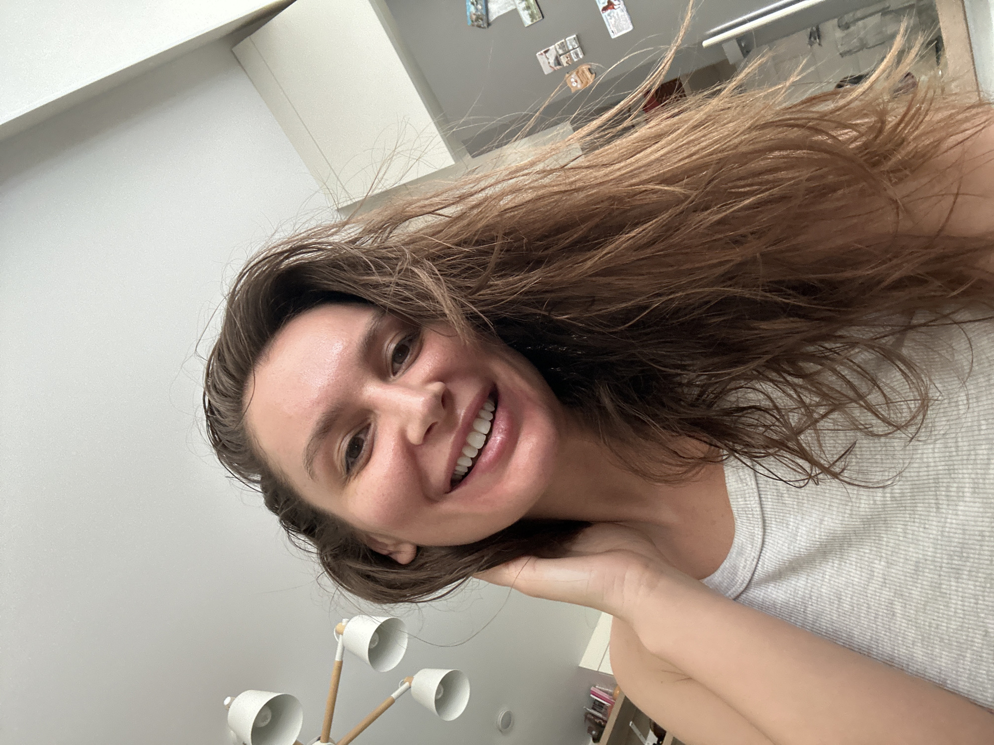 A young woman with medium-long, wavy brown hair is depicted in a tilted, close-up selfie taken indoors, likely in a home setting in Moscow, Russia. Her hair appears somewhat damp or windblown, flowing upwards and to the right in the frame. She is smiling brightly, revealing white teeth, and her expression conveys happiness and cheerfulness. Her left hand is casually placed near her neck or jawline. She is wearing a light-colored, ribbed tank top or sleeveless shirt. In the lower-left foreground, a multi-bulb lamp with white conical shades and light wood-toned accents is partially visible. The background consists of light-colored walls, with a grey surface in the upper right quadrant where several small, rectangular objects, possibly magnets or photos, are attached. The lighting is bright and even, suggesting it is daytime. There is no visible text in the image.