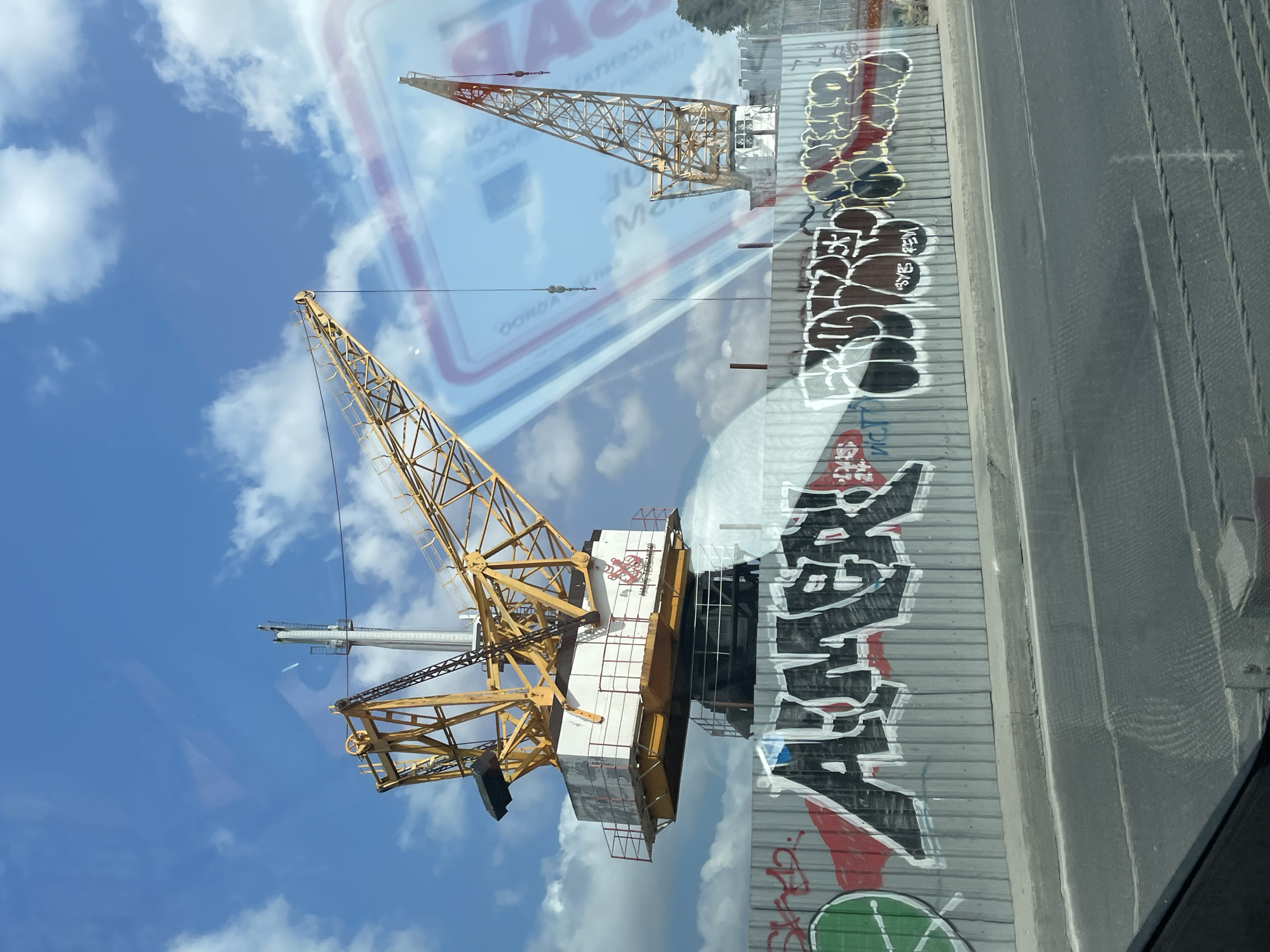 The image shows a construction site with two large yellow cranes against a bright blue sky with white clouds. A corrugated metal fence runs horizontally across the lower half of the image, covered in graffiti. Large black letters that appear to spell "AIMER" are prominently displayed on the fence, along with other graffiti tags in various colors. The cranes are the main focus of the image, with one larger crane on the left and a smaller one on the right. A reflective surface, possibly a window or a car windshield, is visible, showing reflections of clouds and a blurry overlay of what appears to be a credit card or similar object with text. The ground in front of the fence is a paved road with visible tire tracks. The overall scene suggests an urban industrial or construction environment. The presence of construction cranes and graffiti on a fence are common sights in rapidly developing urban areas.