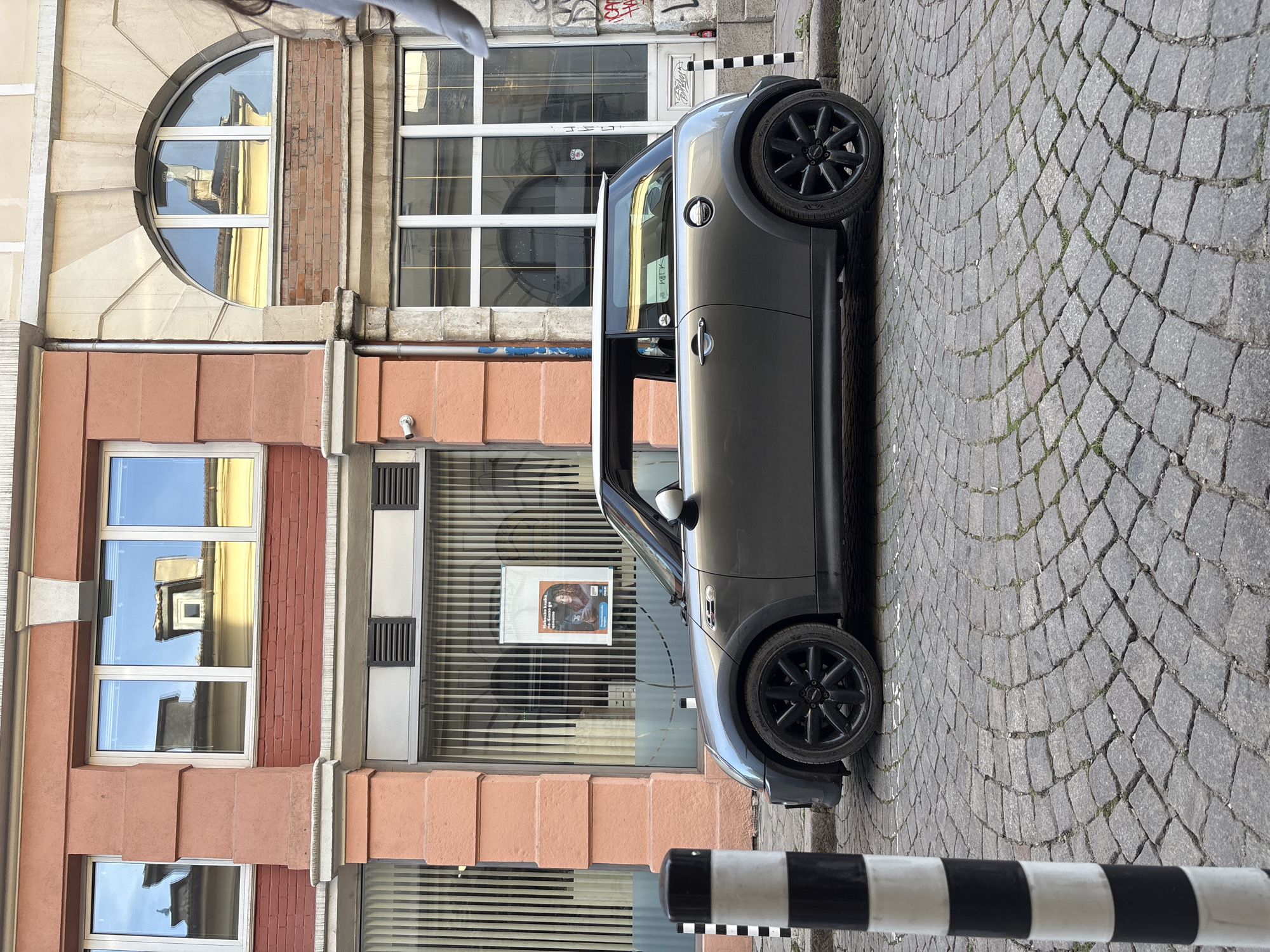 A medium shot depicts a gray Mini Cooper parked on a cobblestone street in Sofia, Bulgaria. The car is parked along a building with a variety of window styles, including a half-circle window on an upper floor and more traditional rectangular windows reflecting nearby buildings. A black-and-white striped bollard is visible near the car's front. A poster is displayed in the building's window behind the car. The environment is urban and well-maintained. No people are visible.