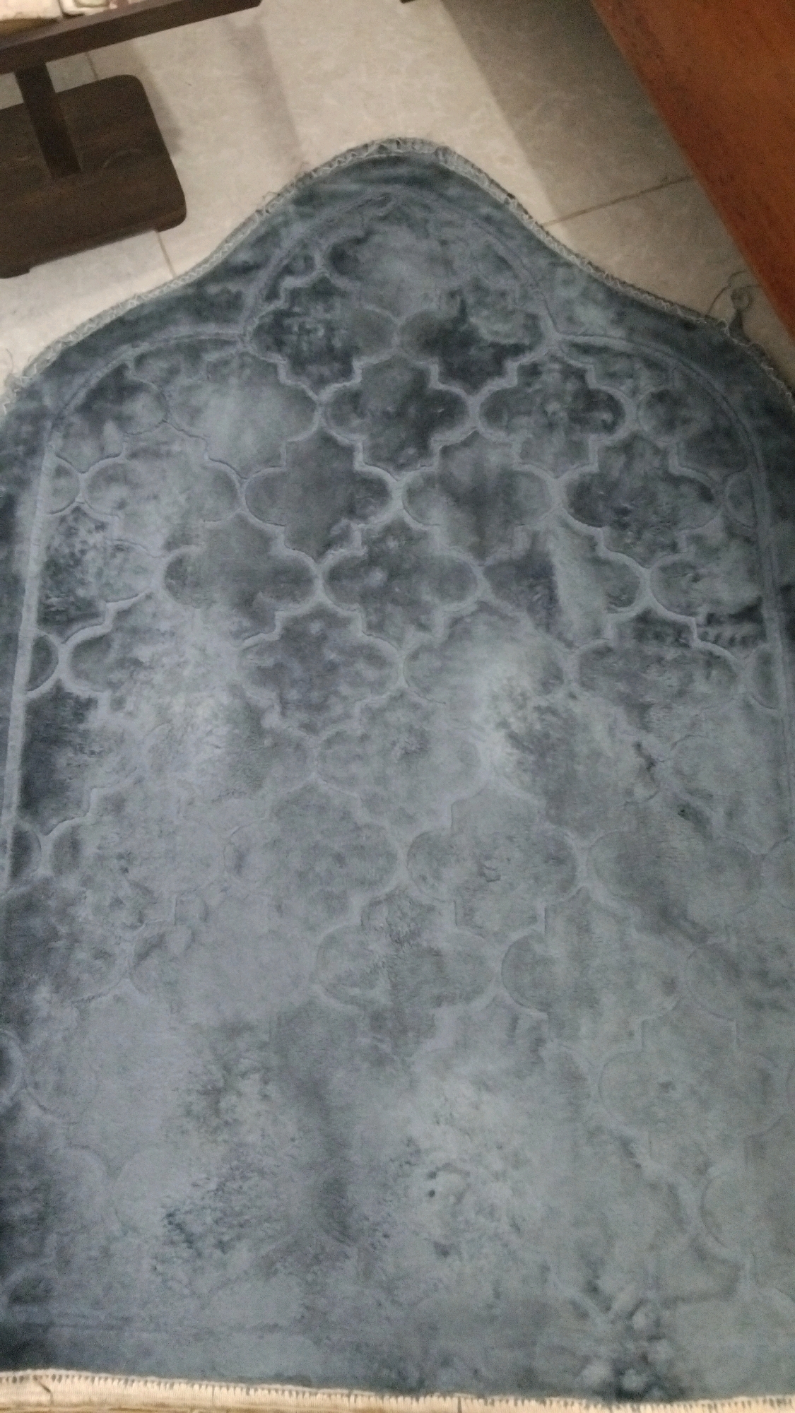 A prayer rug with a quatrefoil pattern is displayed on a tiled floor. The rug is gray and has a fringed edge. A dark wooden table or bench is visible to the left of the rug. The floor is light-colored tile. The image was taken in Kebon IX, Indonesia.