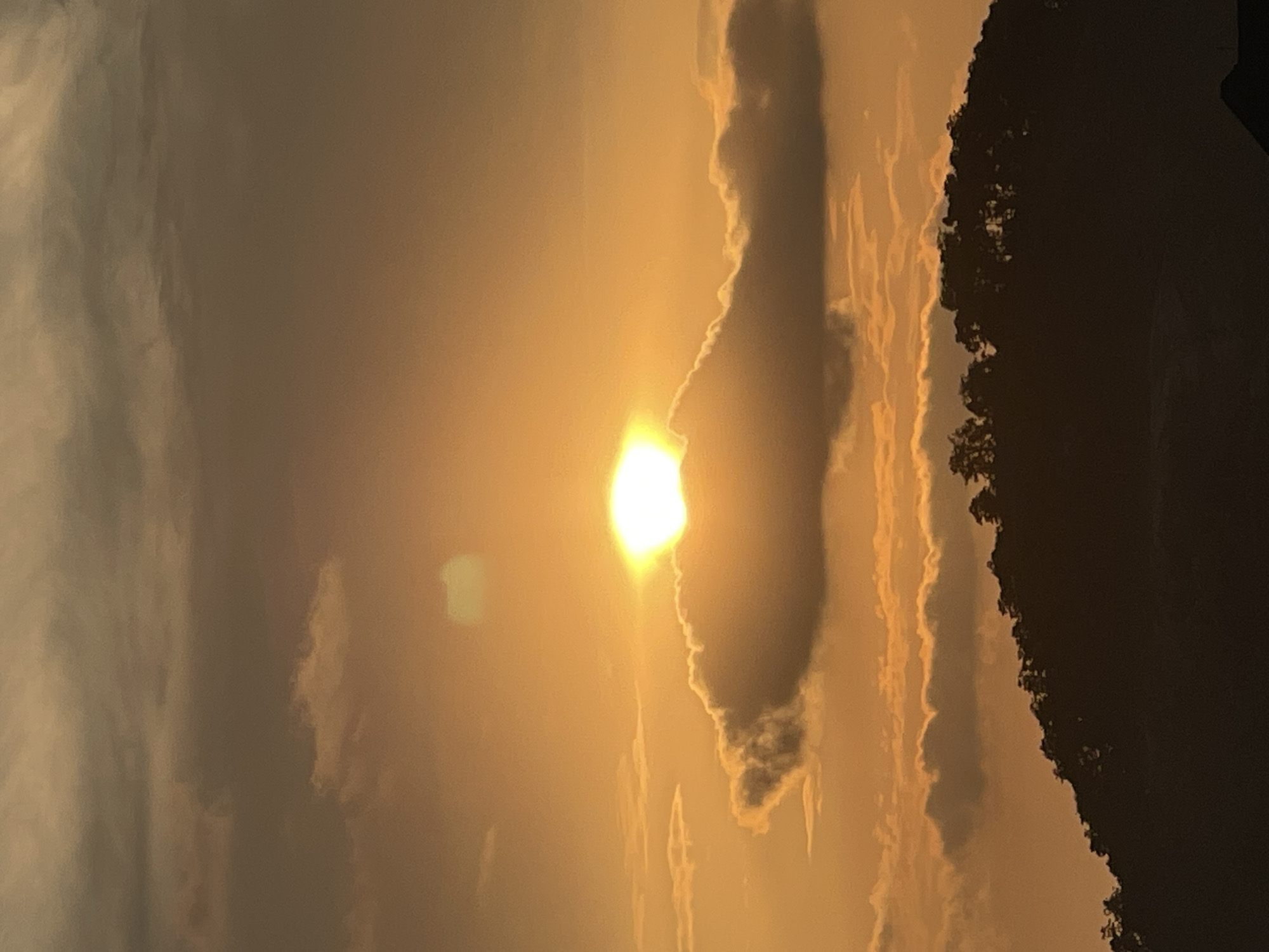 The image shows a sunset over Abuja, Nigeria. The sun is a bright, glowing orb in the sky, partially obscured by a fluffy cloud. The sky is a gradient of orange and yellow, with some darker clouds in the upper portion of the image. Below the sky, a silhouetted treeline of a mountain ridge is visible. The overall impression is one of warmth and tranquility.