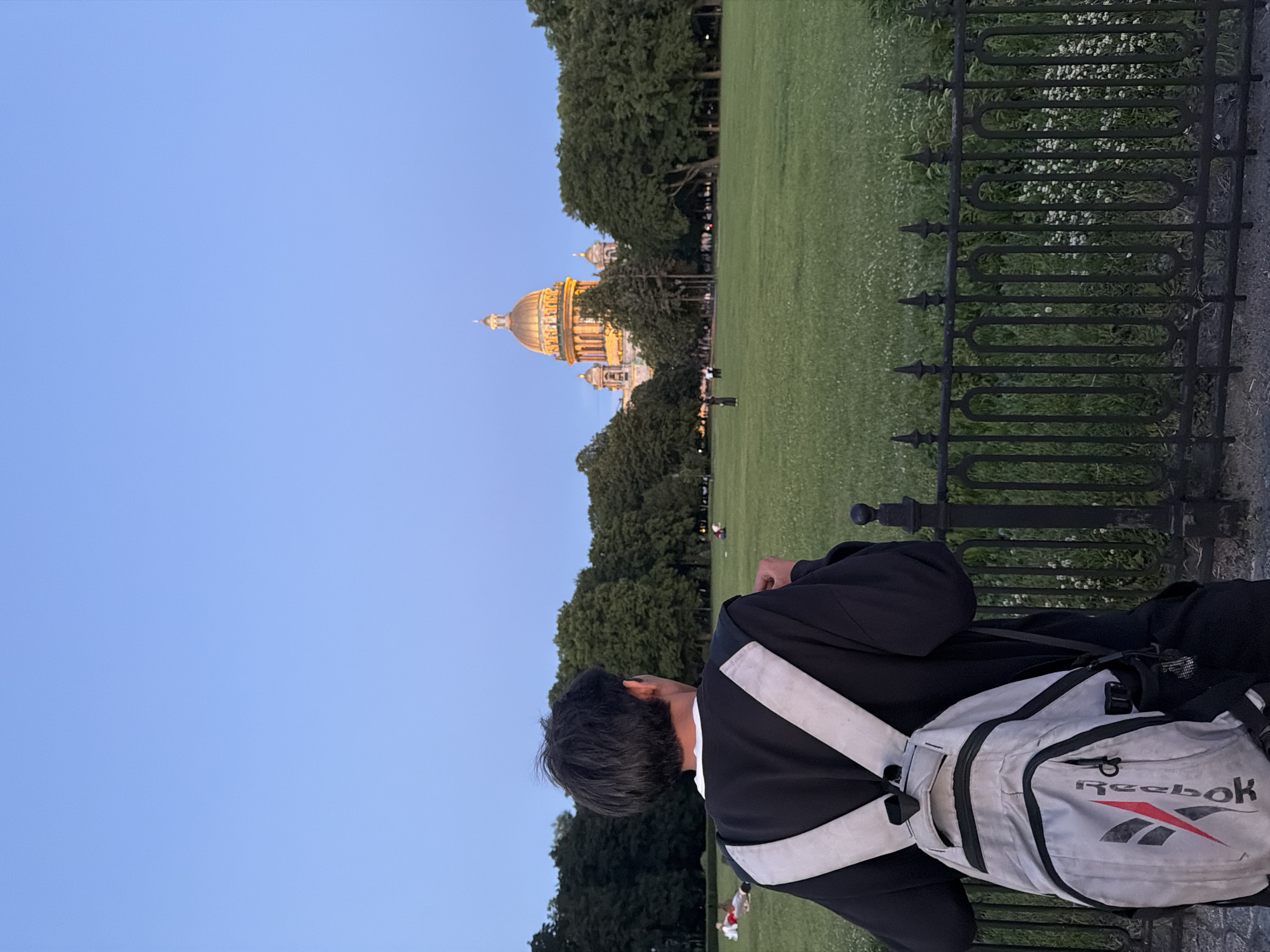 This is a vertical photograph taken outdoors in Saint Petersburg, Russia, likely during the evening or early morning due to the soft, diffused lighting and clear blue sky. The scene depicts a person from behind, wearing a black robe or academic gown with white sashes, and a gray Reebok backpack. They are looking out over a large, grassy park area.
In the distance, a prominent golden-domed building, characteristic of Russian architecture, is visible amongst lush green trees. The building appears to be illuminated, suggesting it is a significant landmark. A dark, ornate metal fence runs along the foreground, separating the viewer's perspective from the park. Small white flowers are scattered in the grass beyond the fence. Several other individuals are scattered across the park, some appearing to be sitting or standing, contributing to a tranquil atmosphere. The overall mood is one of calm observation and appreciation of the cityscape and its architectural heritage. The visible text on the backpack reads "Reebok".